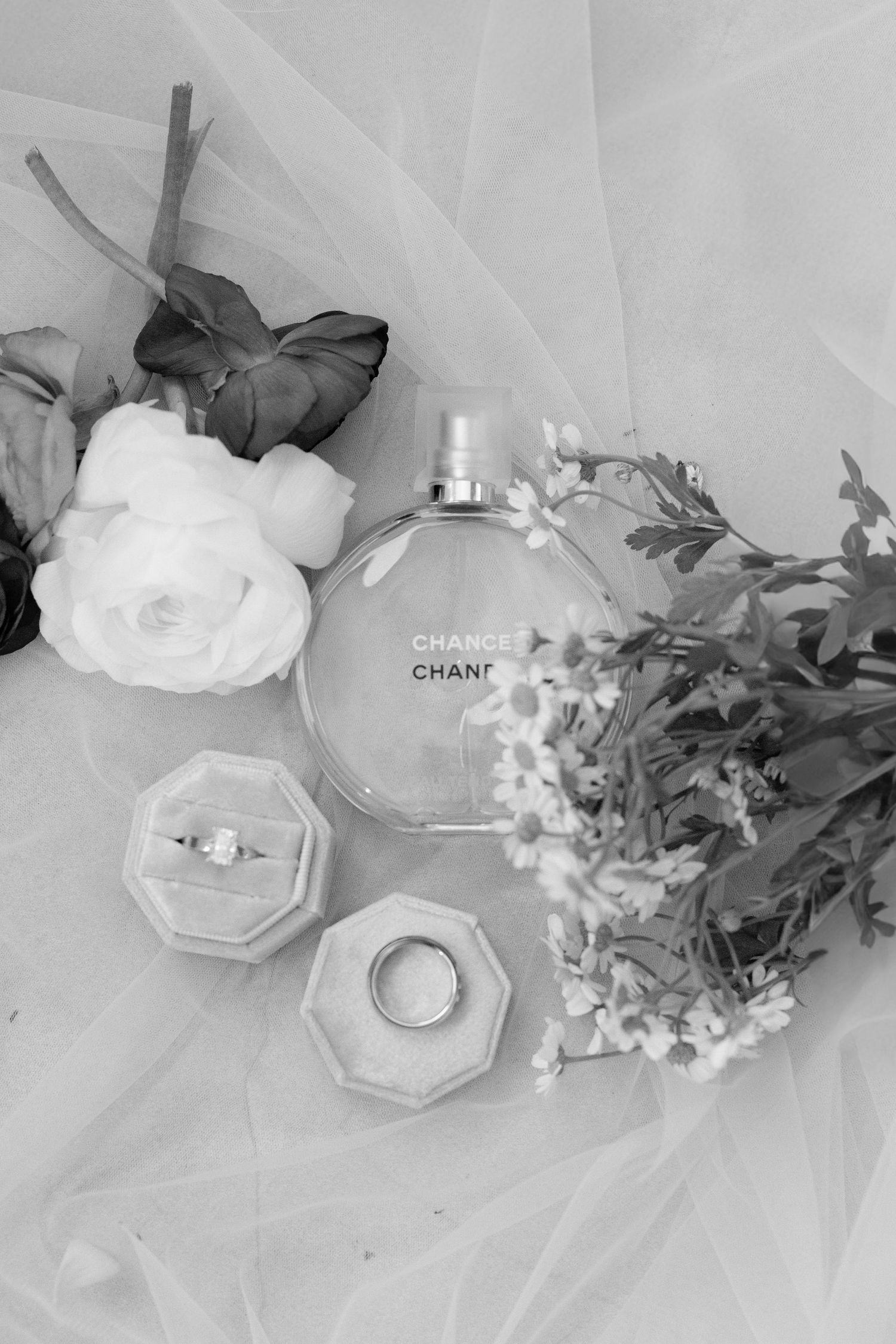 Black and white photograph of a Chanel perfume bottle, ring boxes, and delicate flowers on tulle fabric.