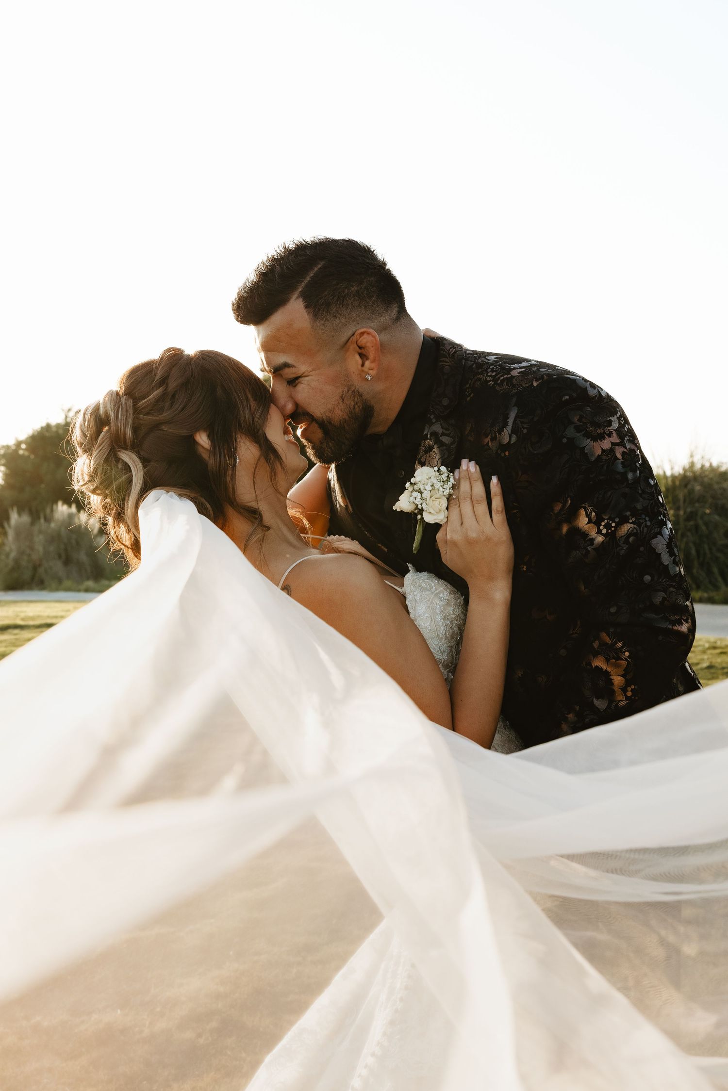 A romantic wedding photo at sunset with the bride's veil flowing dramatically in the wind as the couple shares an intimate moment.