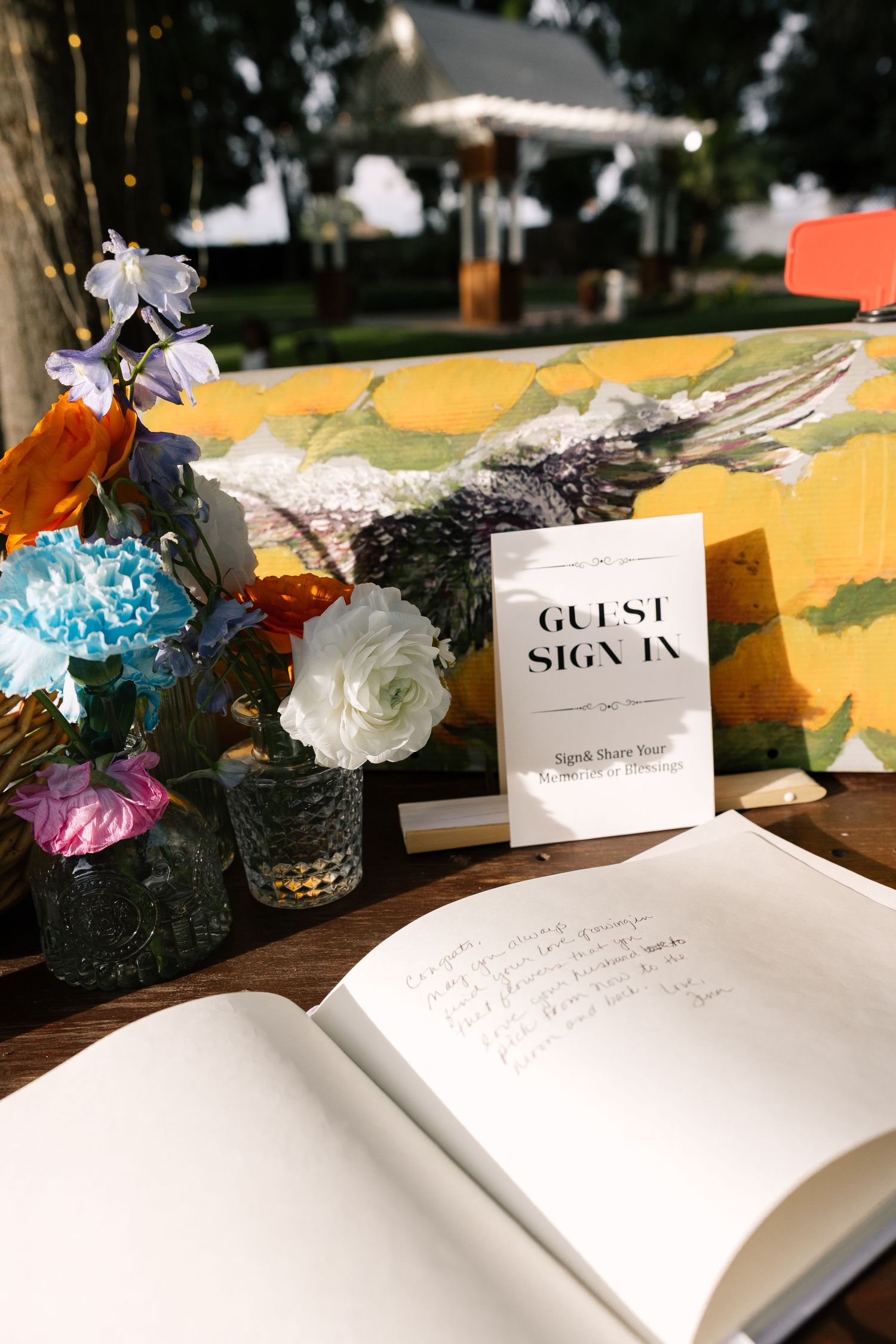 An open guest sign-in book with colorful flowers in vases and a sign displayed on a rustic wooden table outdoors.