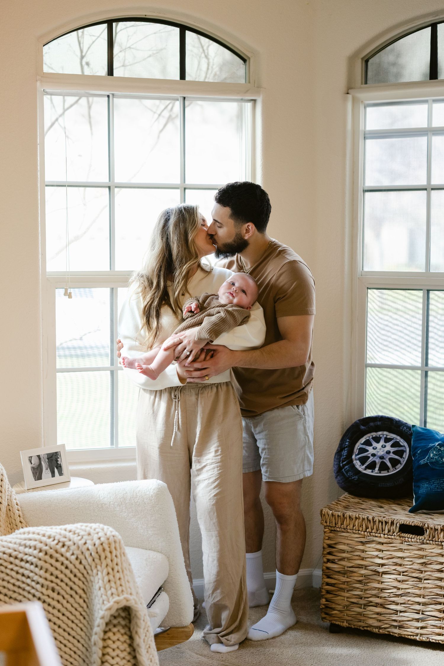 A couple shares an intimate moment near bright windows while holding their newborn baby in a cozy home setting.