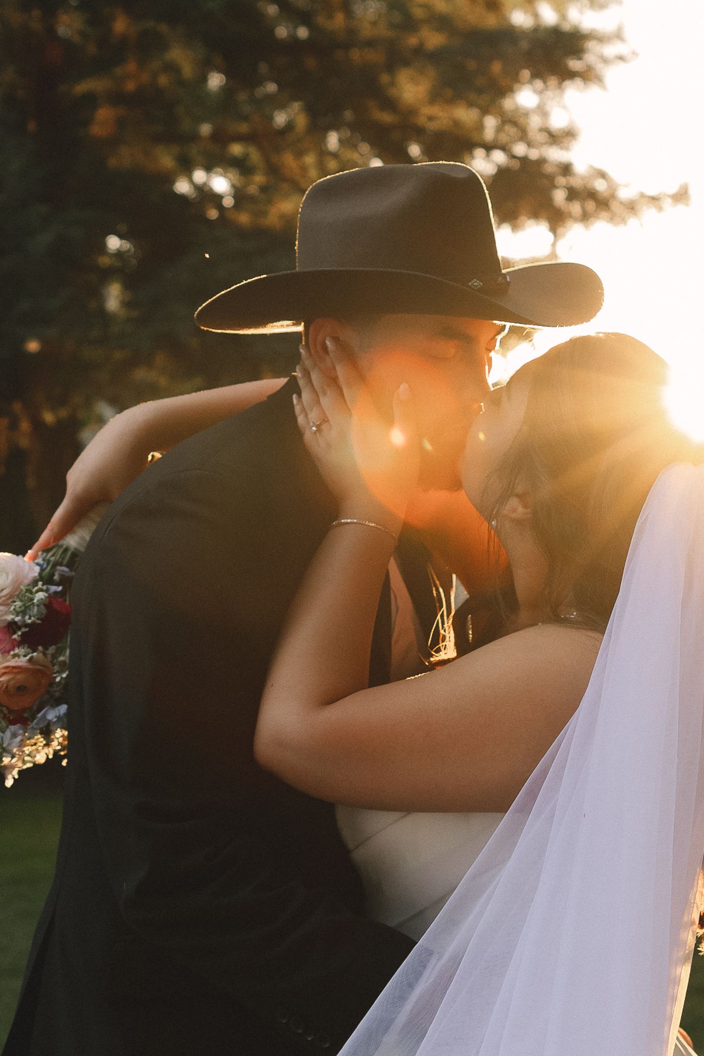 A romantic wedding photo at sunset showing a couple embracing, with the groom wearing a cowboy hat and the bride in a veil.