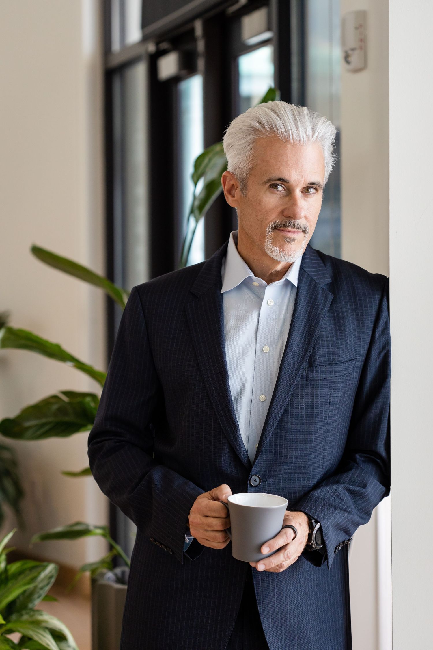 Mature businessman with gray hair holding coffee cup, wearing navy suit, standing by window with indoor plants.