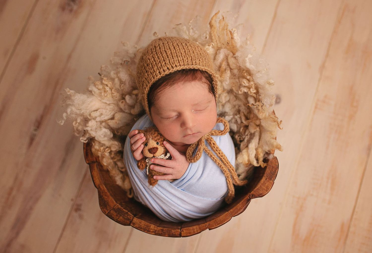 Sleeping newborn wearing knit hat and blue swaddle lies in wooden bowl with soft dried flowers on wood floor.