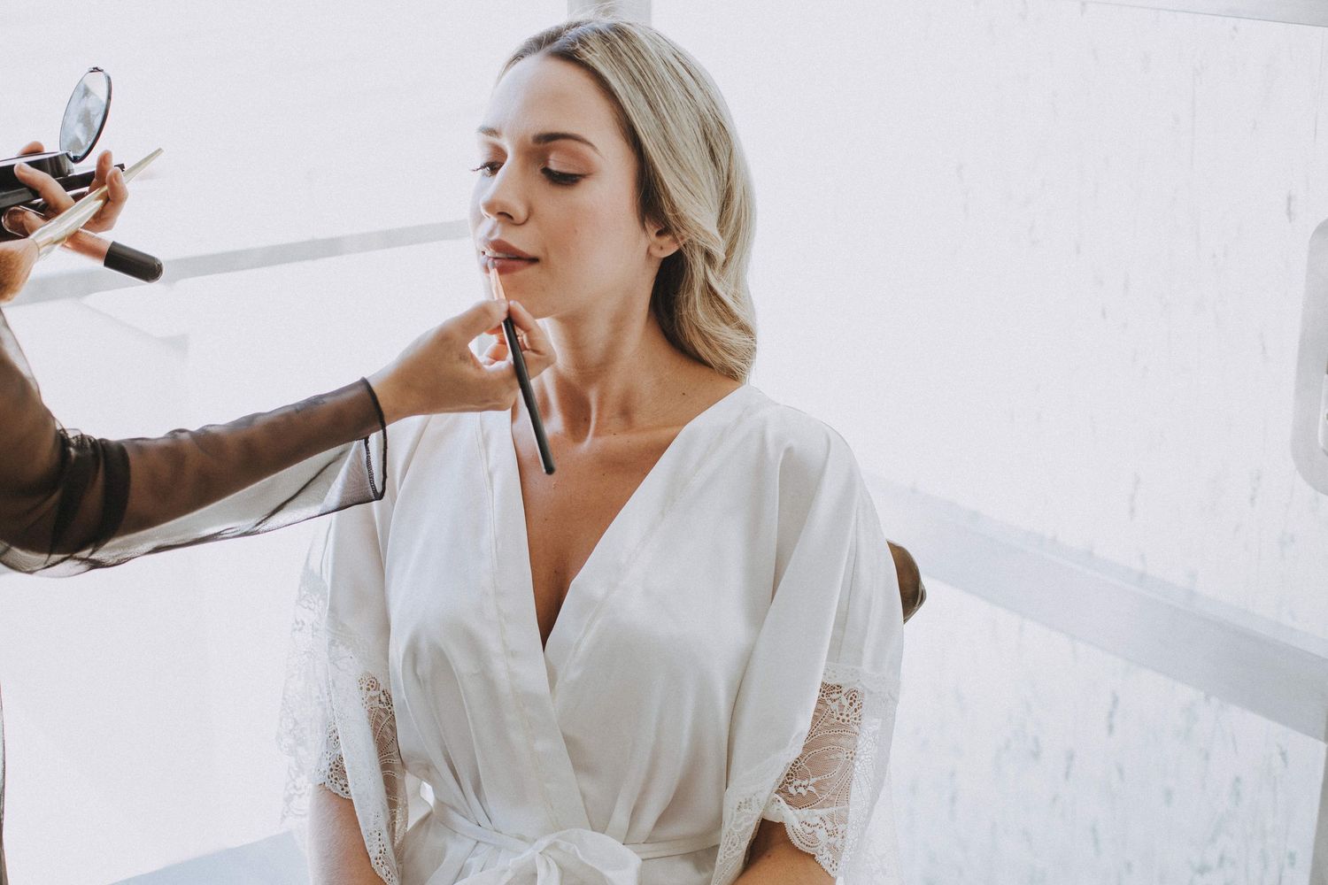 A bride getting ready in a white robe sits while having her hair and makeup professionally styled.
