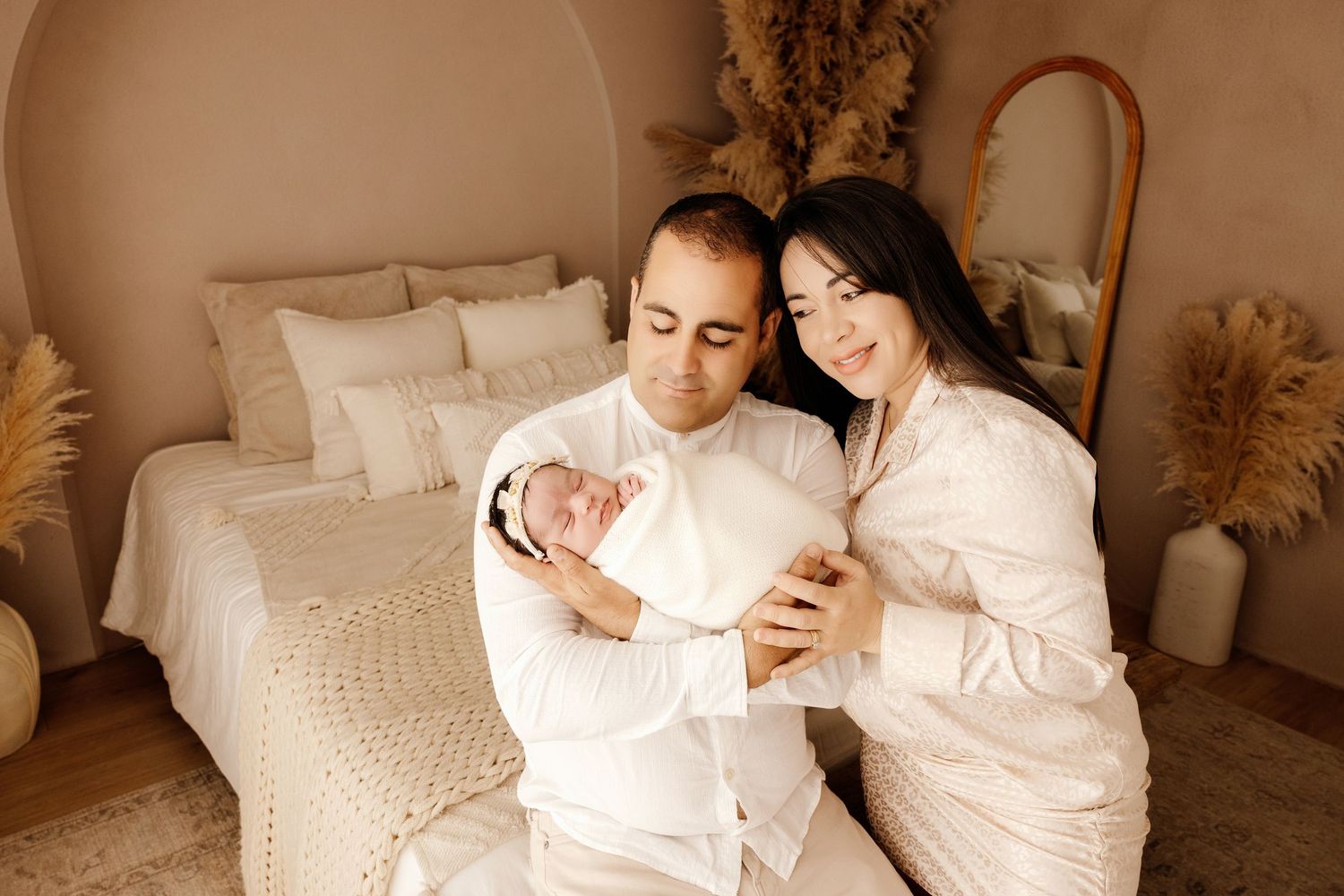 A couple in white outfits tenderly holds their newborn baby in a cozy, neutral-toned bedroom setting.