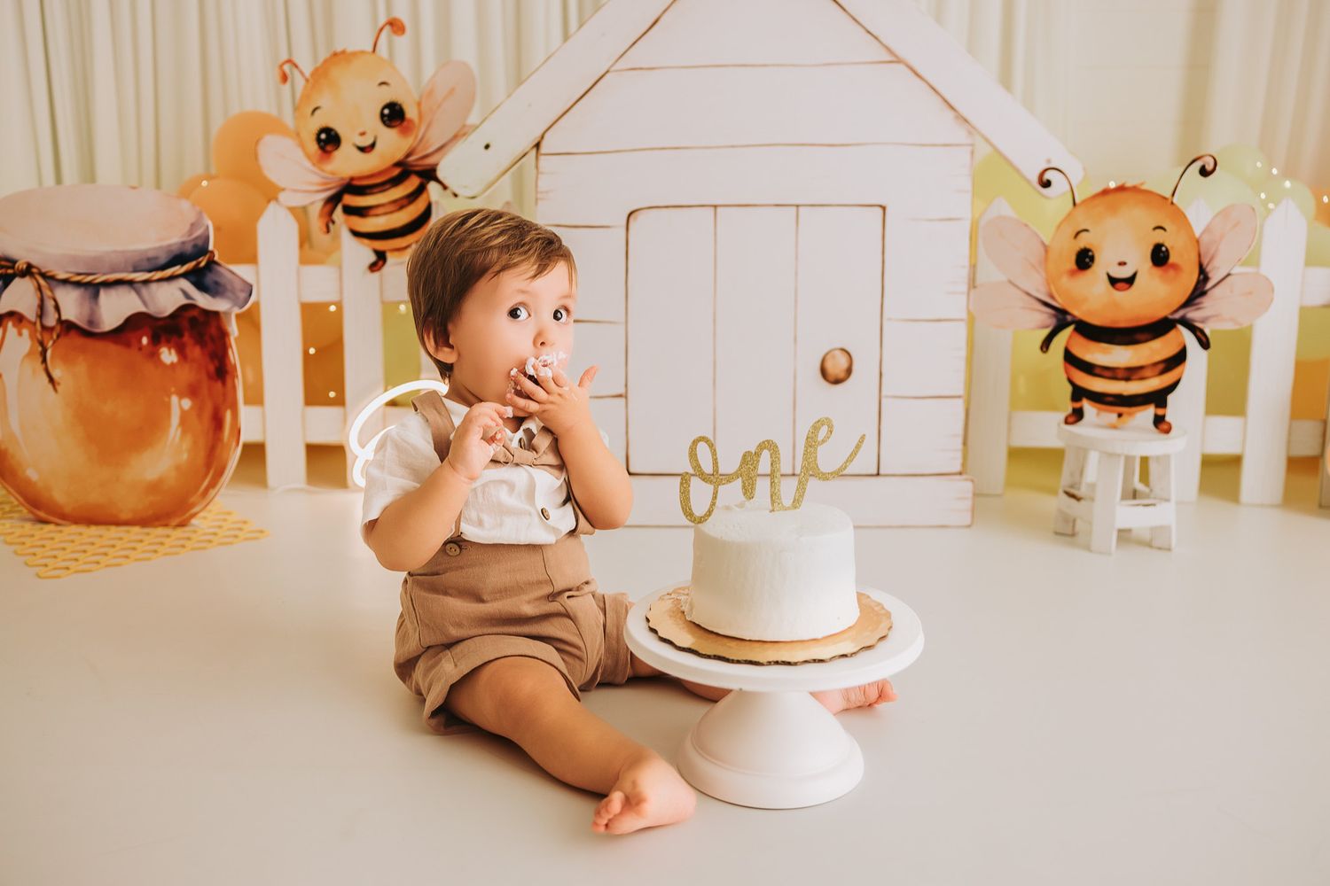 Baby boy smashing first birthday cake surrounded by bee-themed decorations and a white playhouse backdrop.