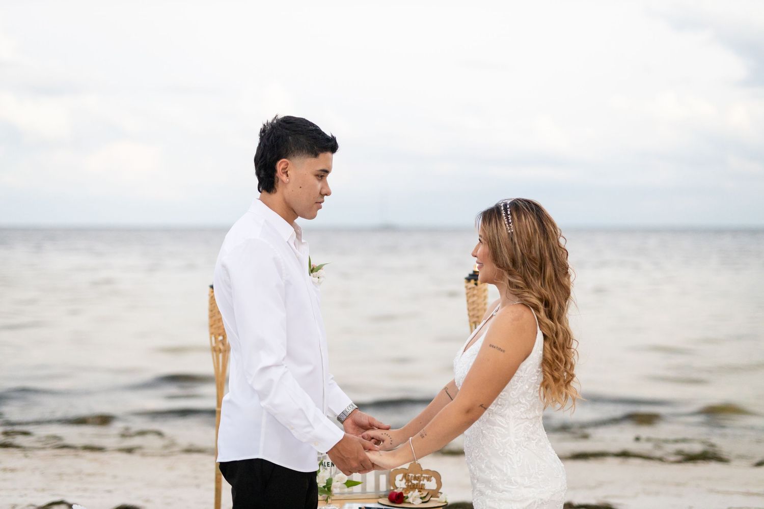 A couple in white outfits holds hands while walking along a scenic beach on an overcast day.