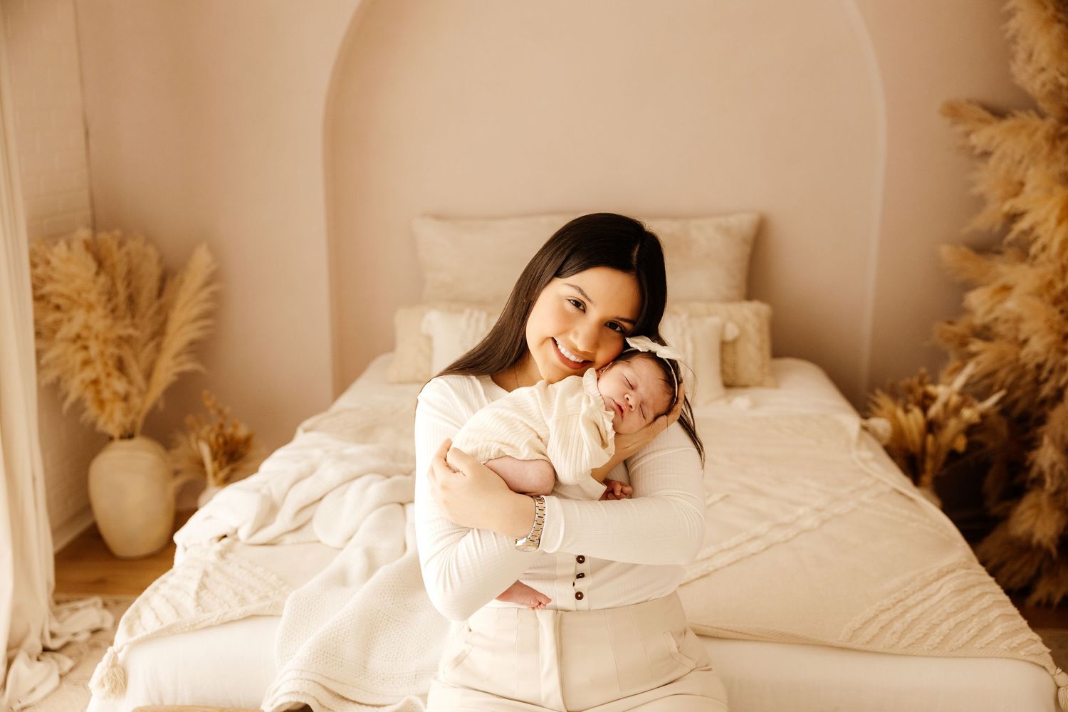 A mother in a white dress tenderly holds her sleeping newborn baby while sitting in front of a bed.