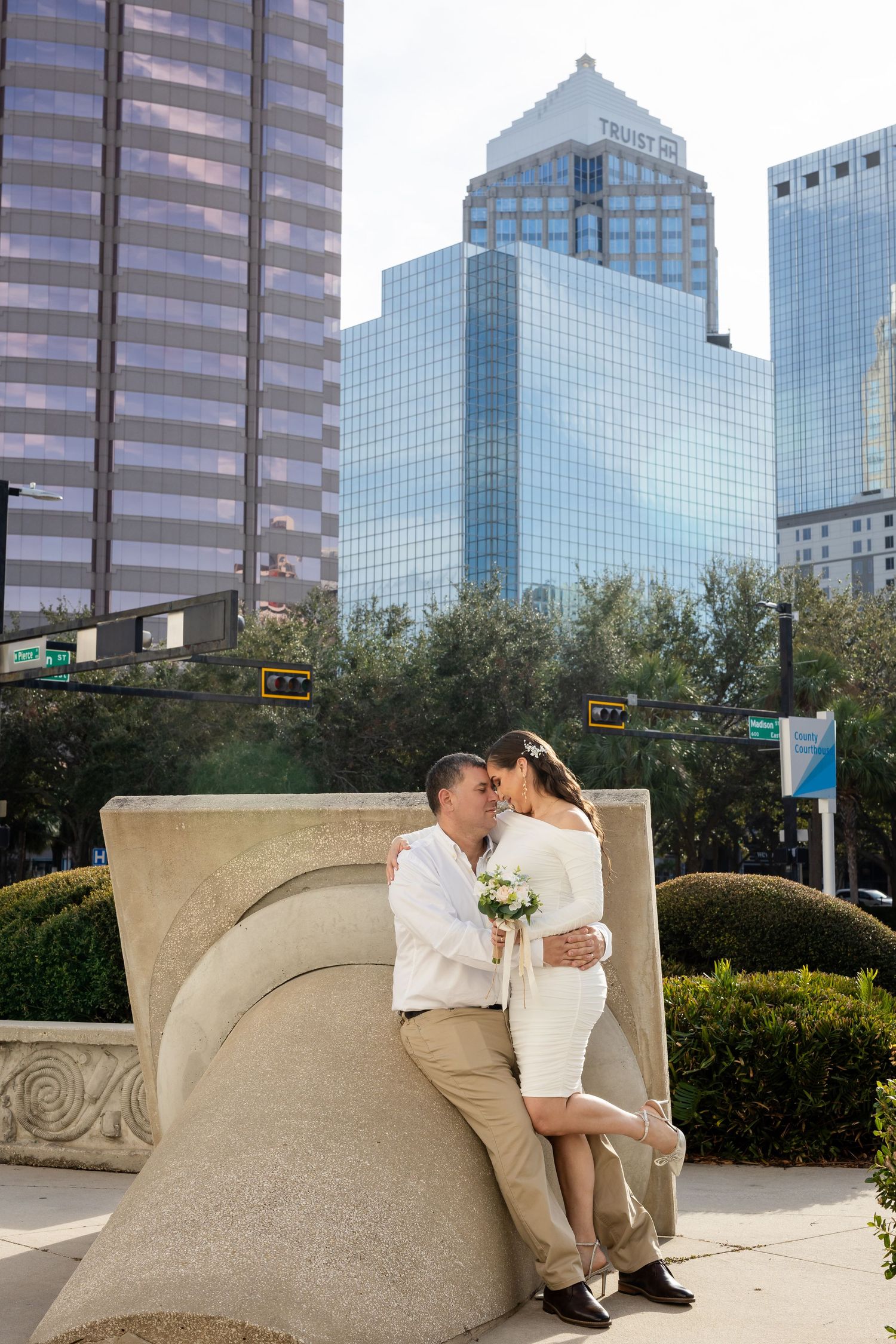 Romantic couple sitting on sand-colored curved bench with modern glass buildings in urban skyline background.