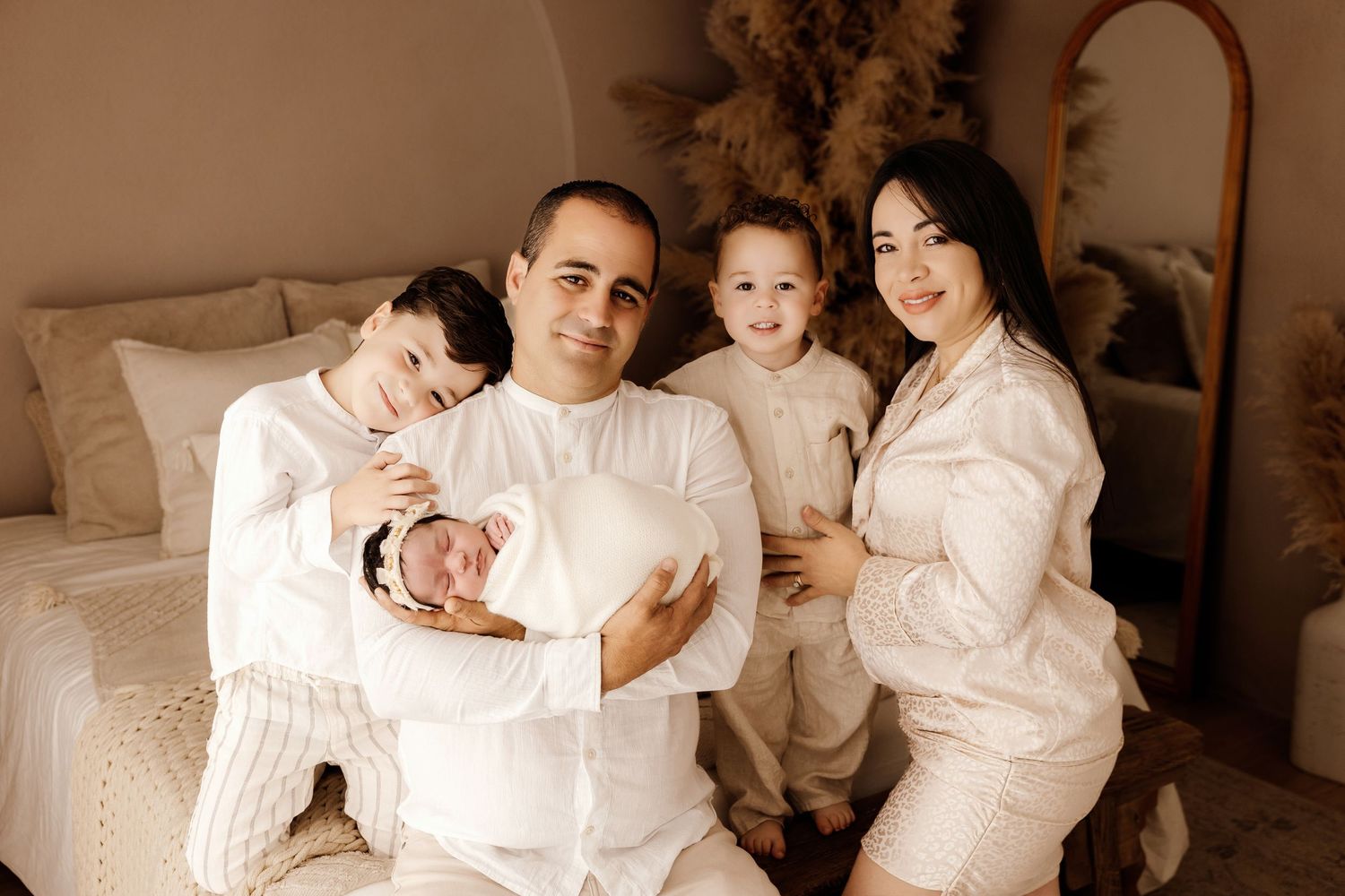 Family of four in matching white outfits posing together, parents holding newborn baby and toddler in cozy bedroom setting.