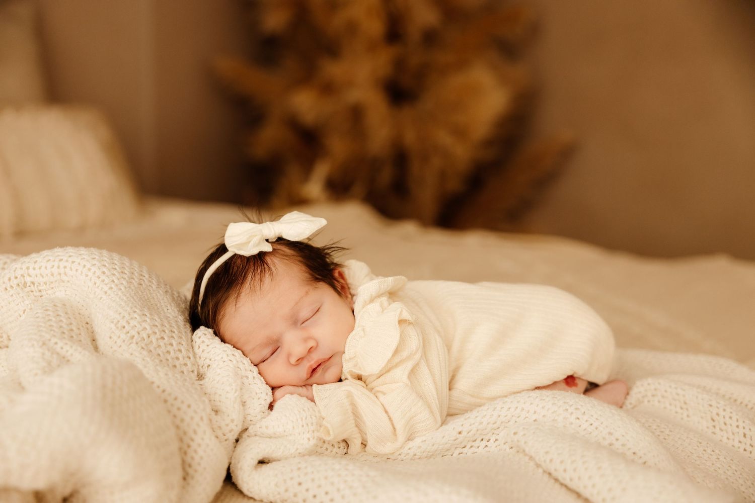 Peaceful newborn baby sleeping on white knit blanket wearing cream outfit and bow headband.