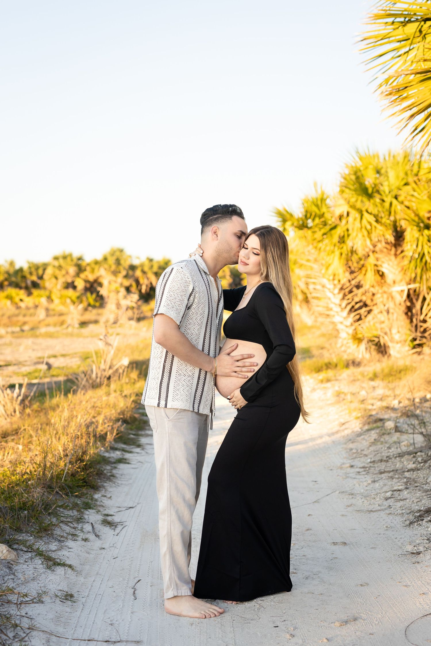Expecting couple embraces on sandy beach path surrounded by palm trees at golden hour.