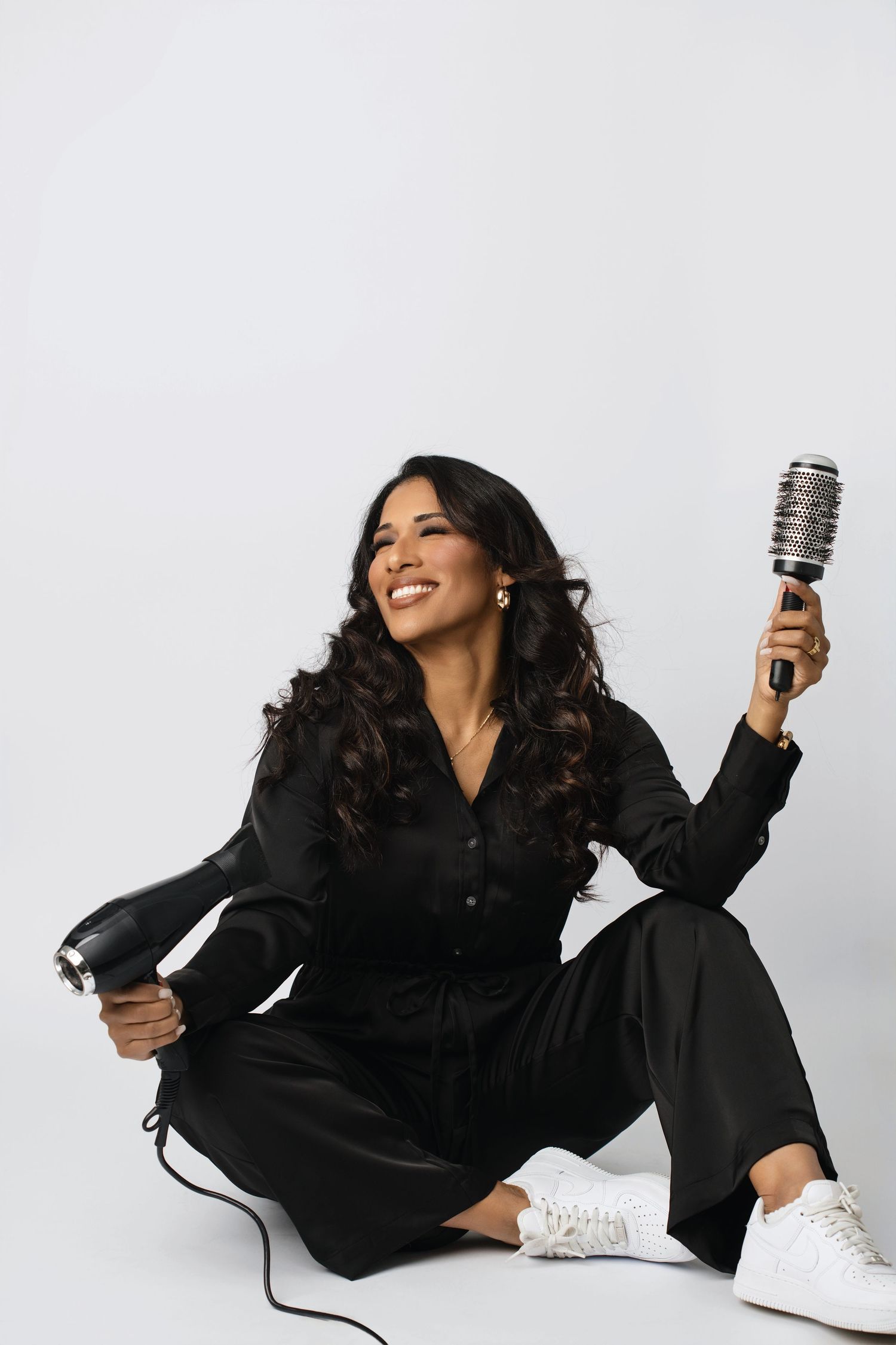 Smiling woman in black outfit sitting on floor holding a hair dryer and round brush on white background.