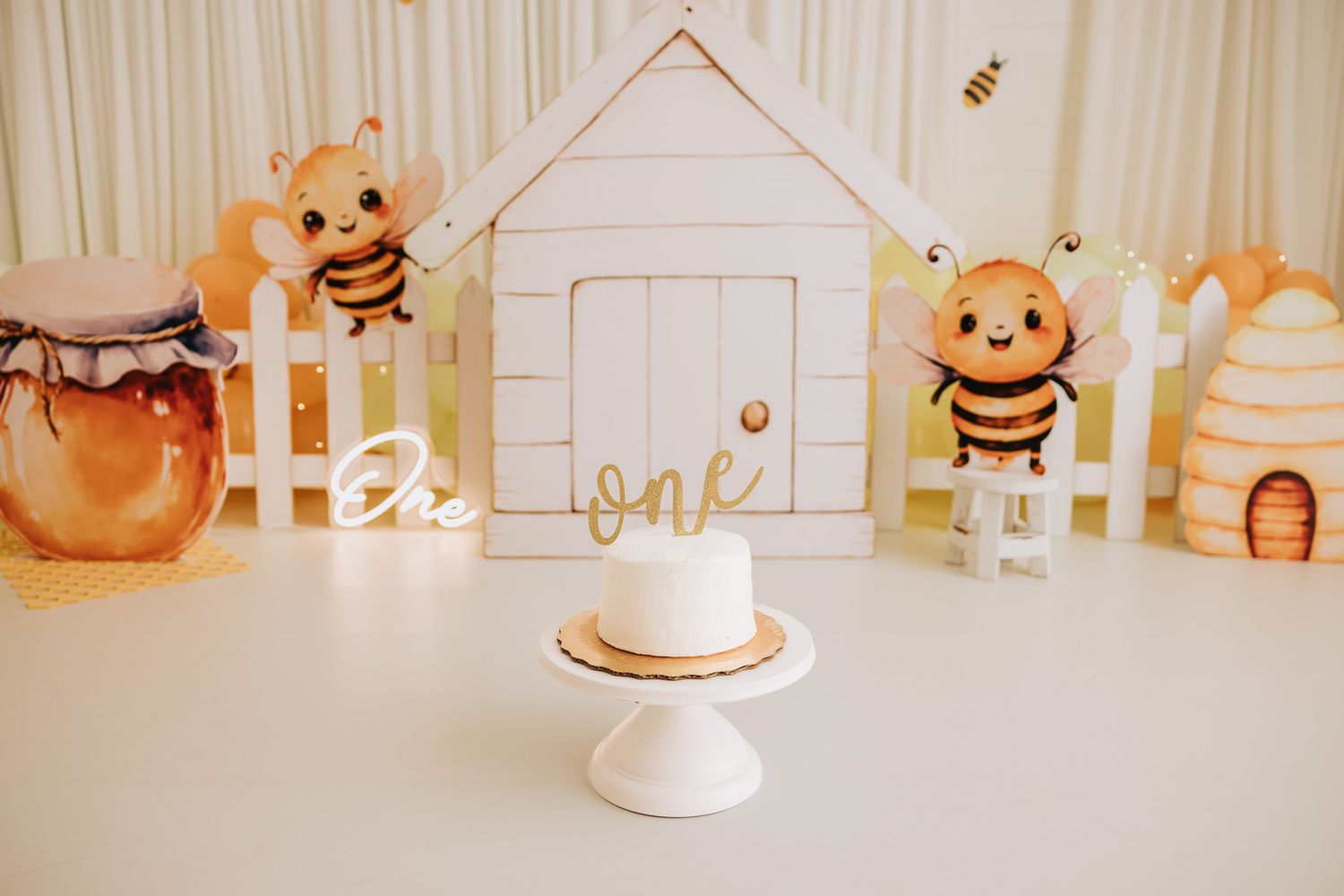 Bee-themed first birthday cake smash setup with balloon bees, honeycomb decor, and a white cake reading One.