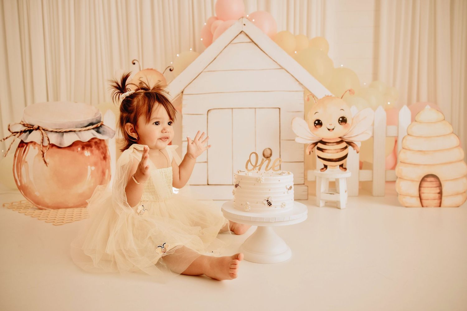 Baby girl in white dress sits by a small cake at a bee-themed first birthday smash cake photo session.