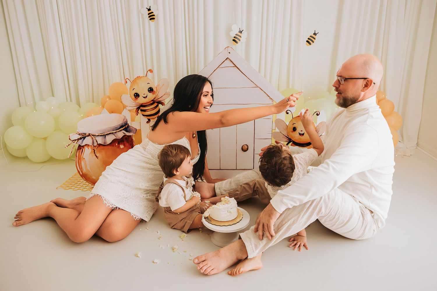 Family celebrating baby first birthday with bee theme, balloons, and cake in a bright white studio setting.