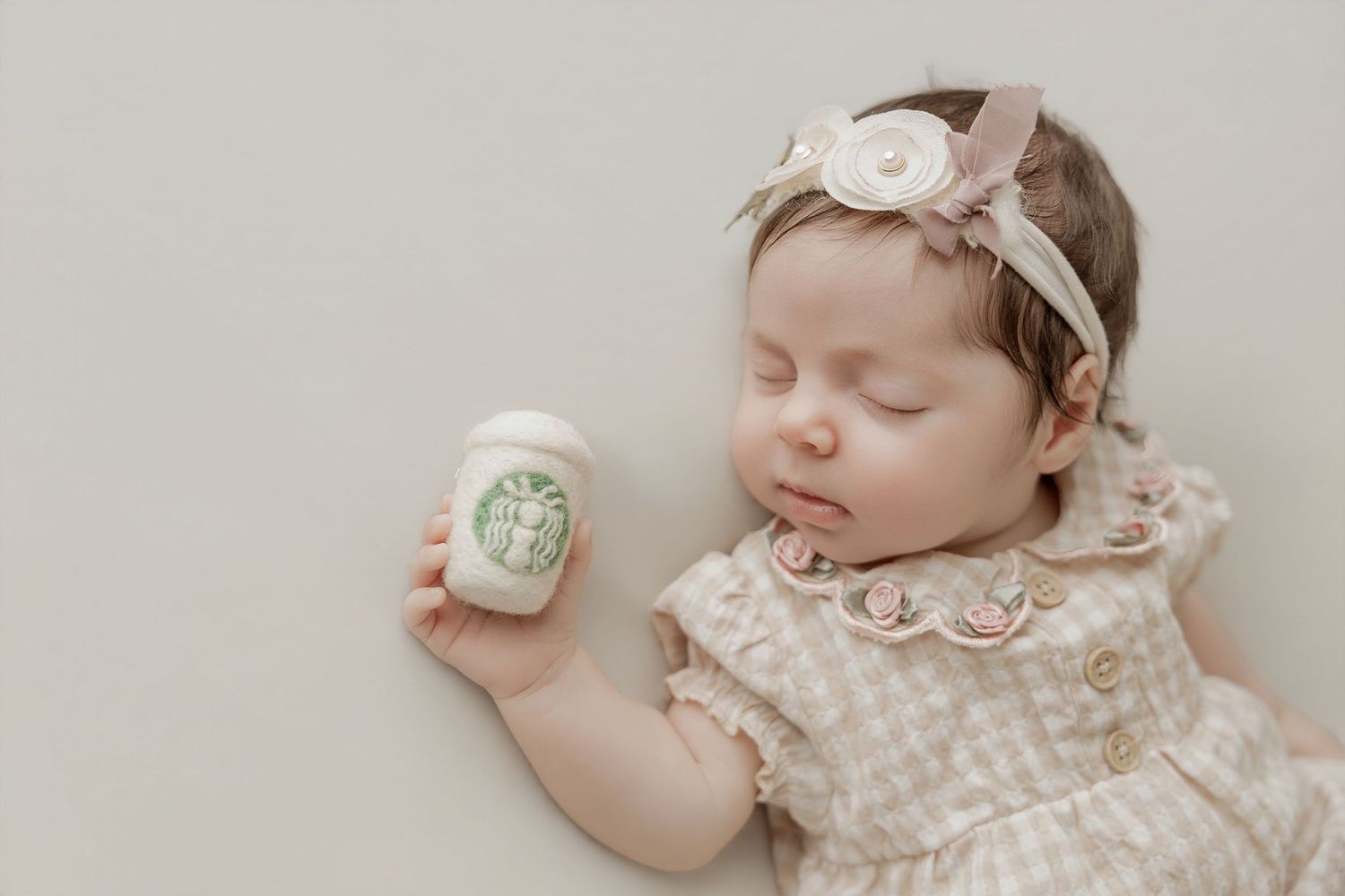 Sleeping baby in beige outfit holding mini Starbucks cup toy, wearing floral headband on neutral background.
