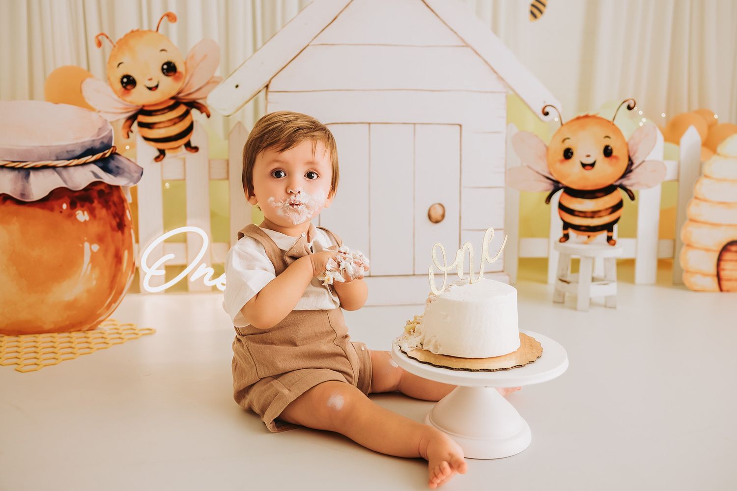 Cute baby boy enjoys first birthday cake smash surrounded by bee-themed balloons and decorations.