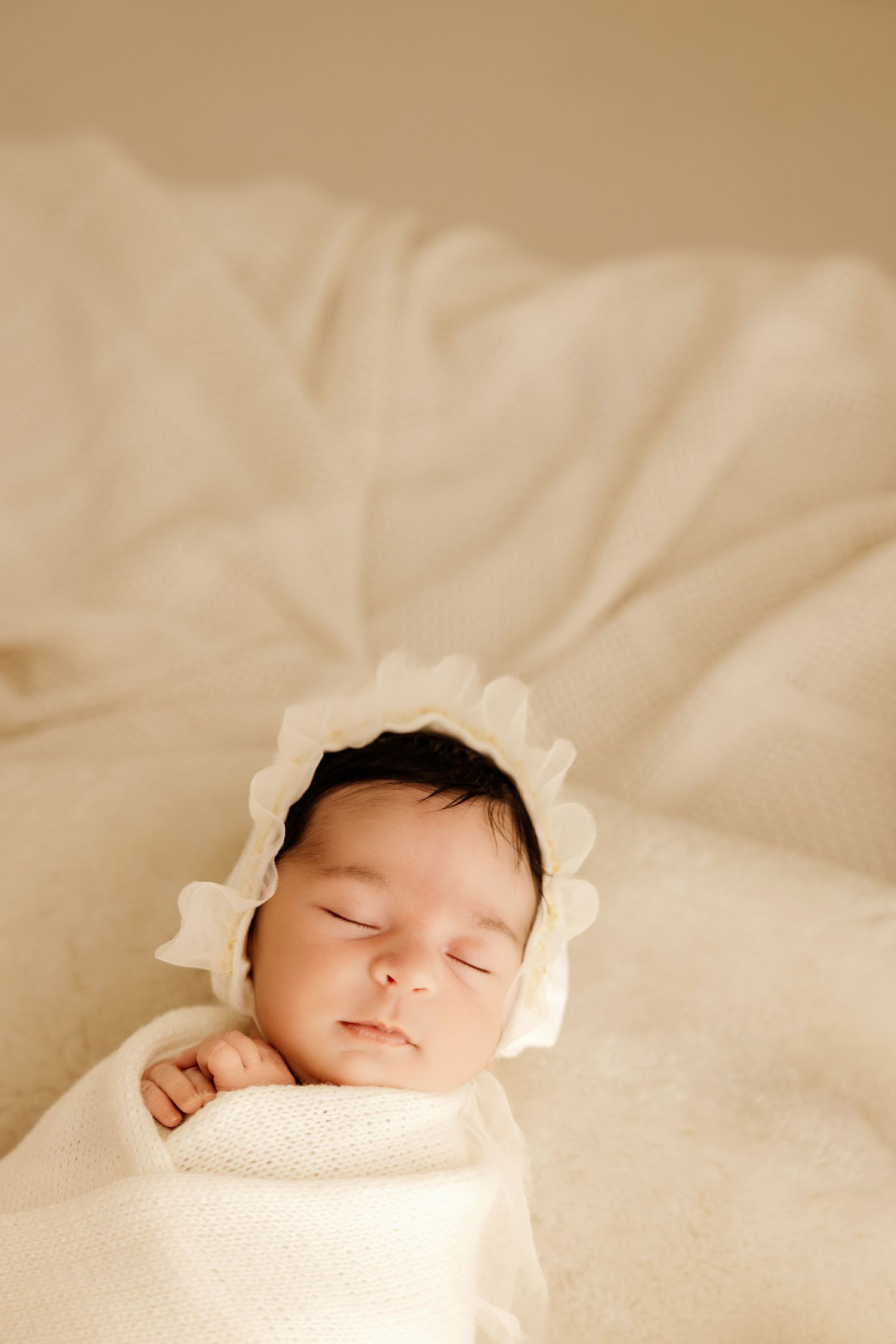 Peaceful newborn baby sleeping wrapped in cream knit blanket wearing white lace bonnet, soft warm tones.
