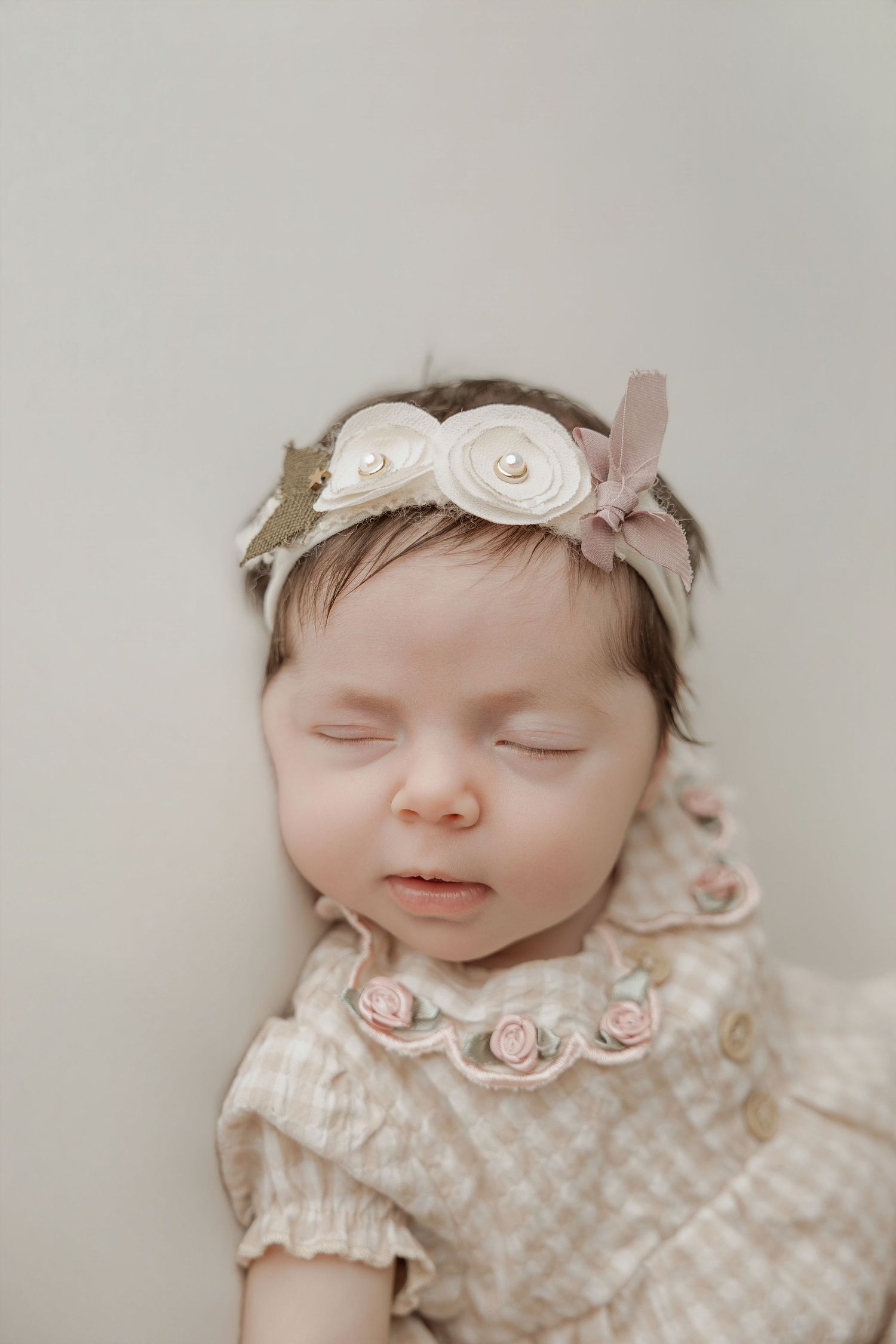 Sleeping newborn baby girl wearing a floral headband and embroidered rose outfit against a neutral background.