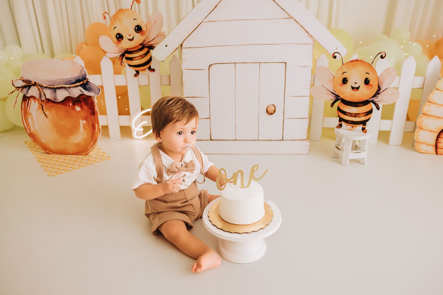 Baby boy sits by smash cake at bee-themed first birthday party with honey jar decorations and cardboard house backdrop.