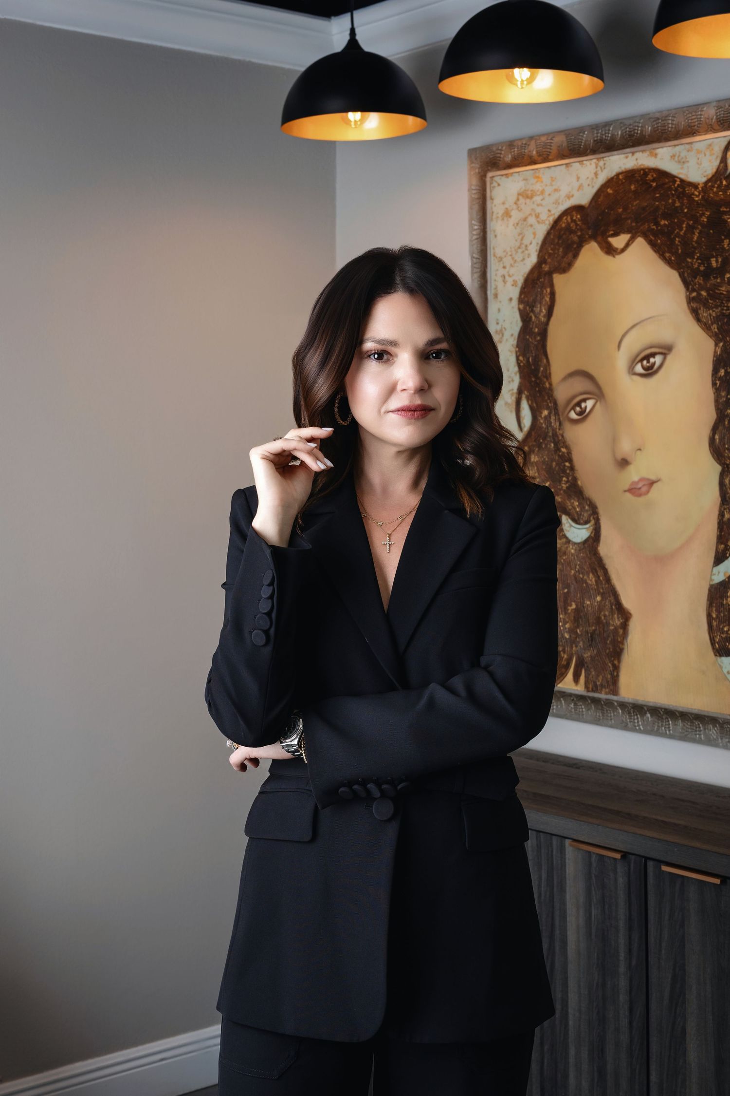 Professional woman in black blazer suit posing confidently in modern office with decorative portrait painting.