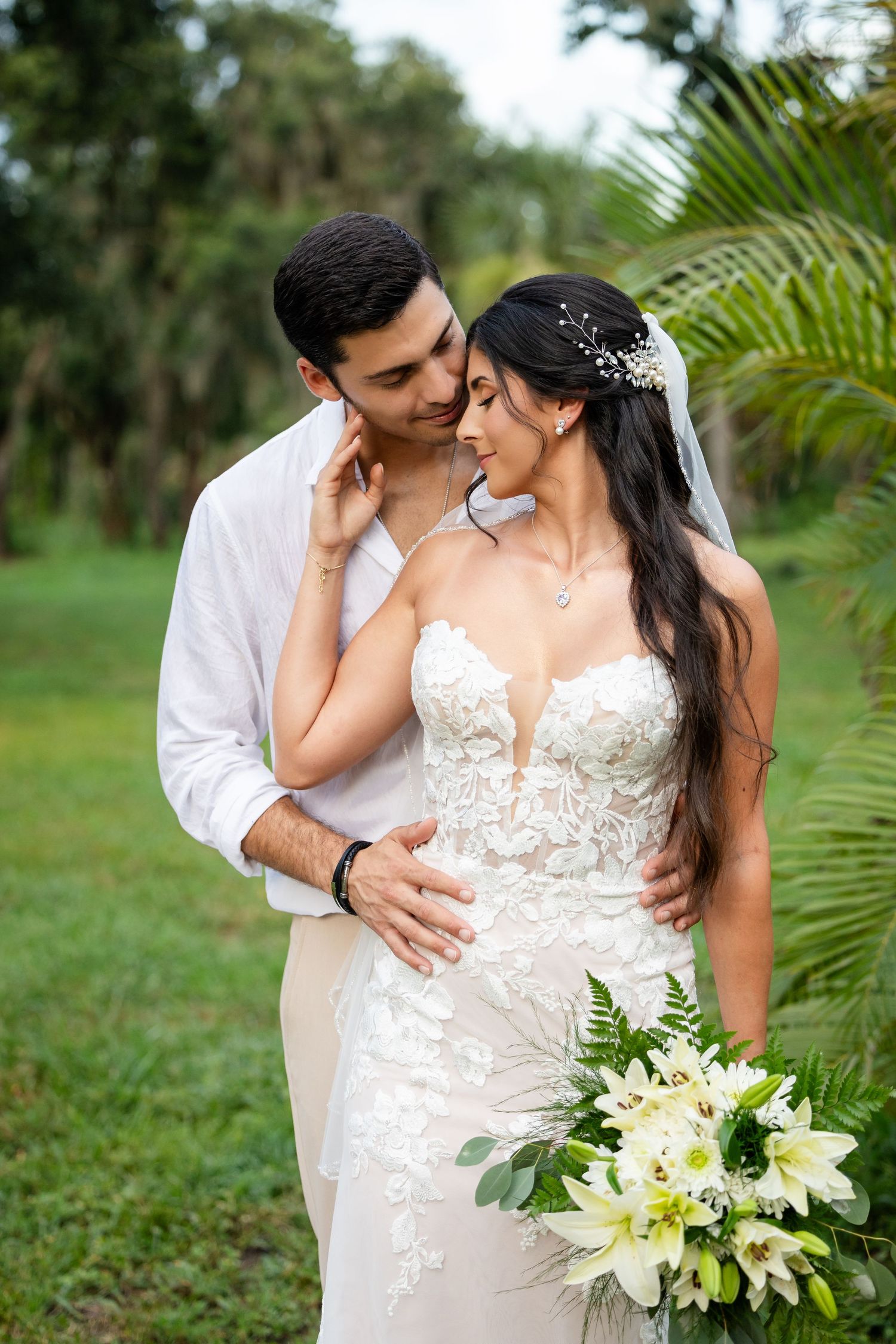 Romantic outdoor wedding photos with couple embracing among tropical palms and white lily bouquet.