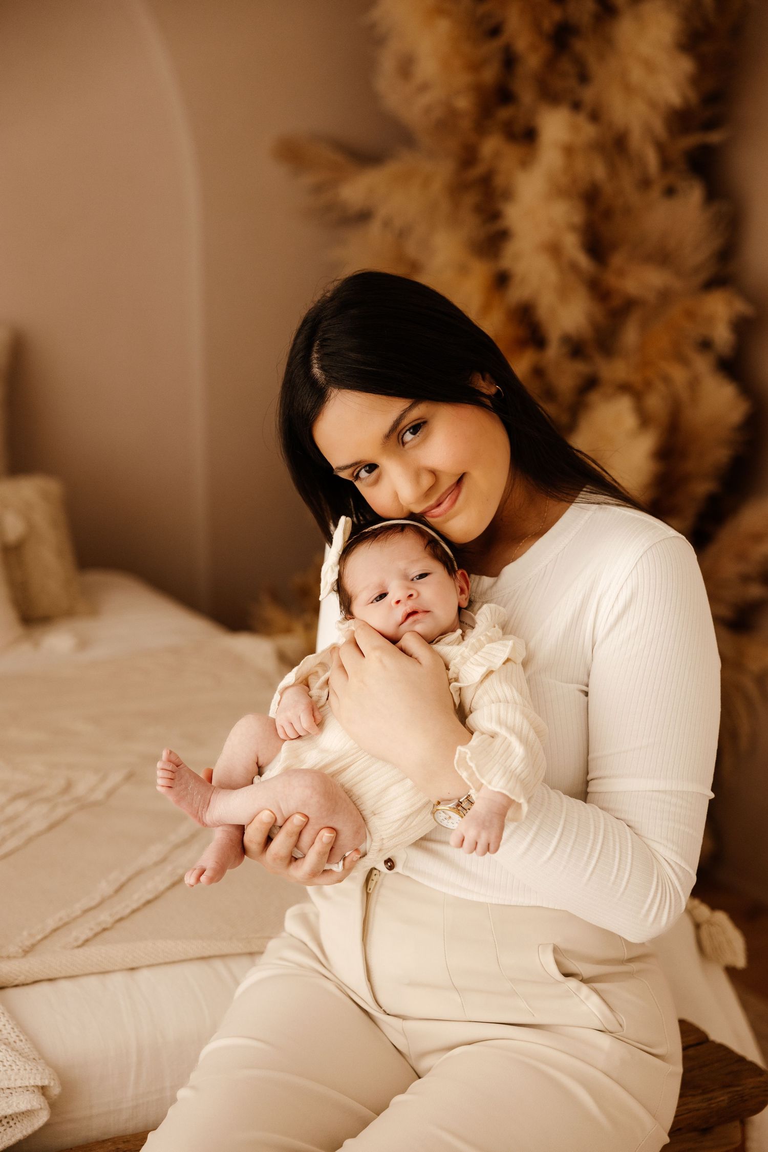 A smiling dark-haired woman in white outfit holds her newborn baby in a warm beige boho-styled bedroom.