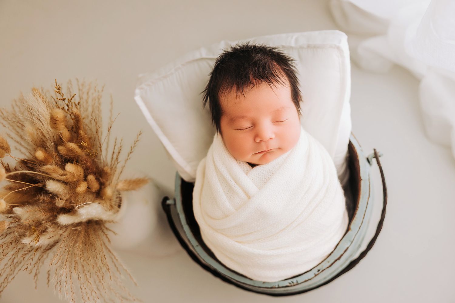 Newborn baby swaddled in white wrap sleeping peacefully in a wooden bucket beside dried pampas grass.