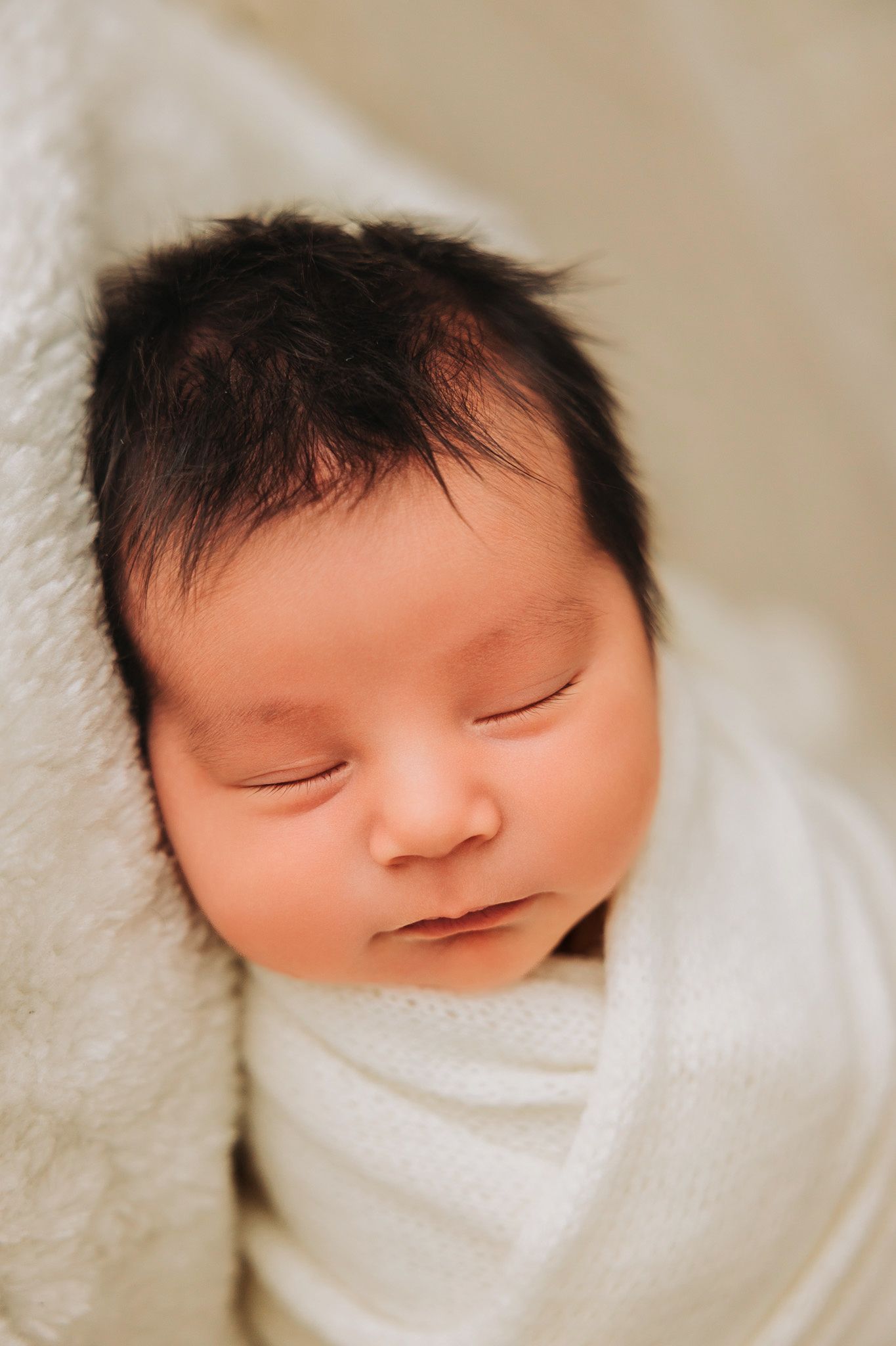 Peaceful newborn baby with dark hair sleeping wrapped in a soft white swaddle blanket.