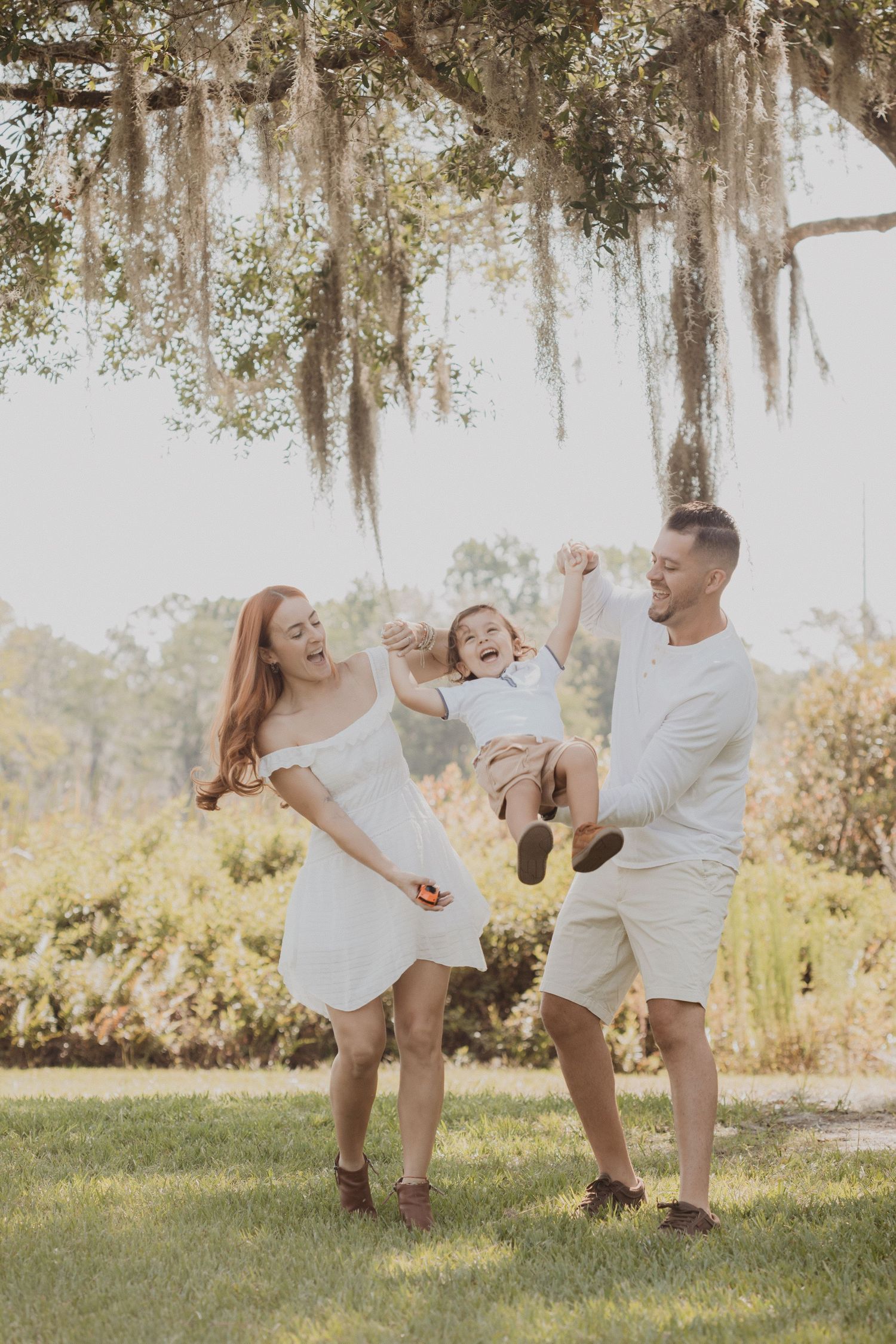 Family in white outfits plays and laughs together under Spanish moss in a sunlit garden setting.