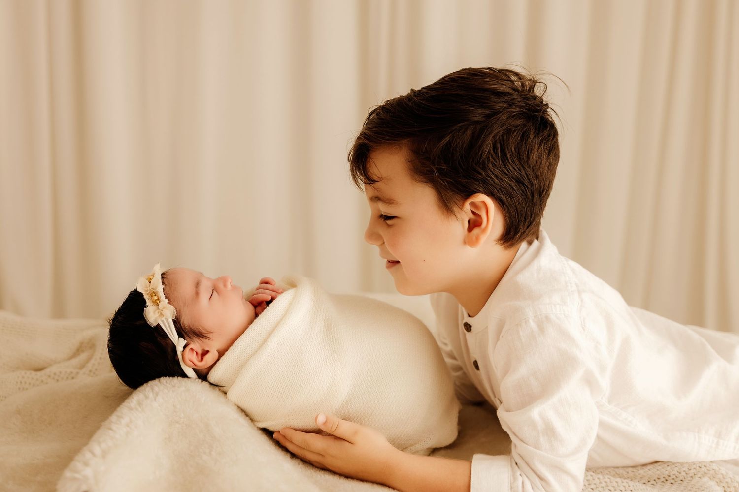 A young boy in white shirt tenderly gazes at a swaddled newborn baby lying on a soft blanket.