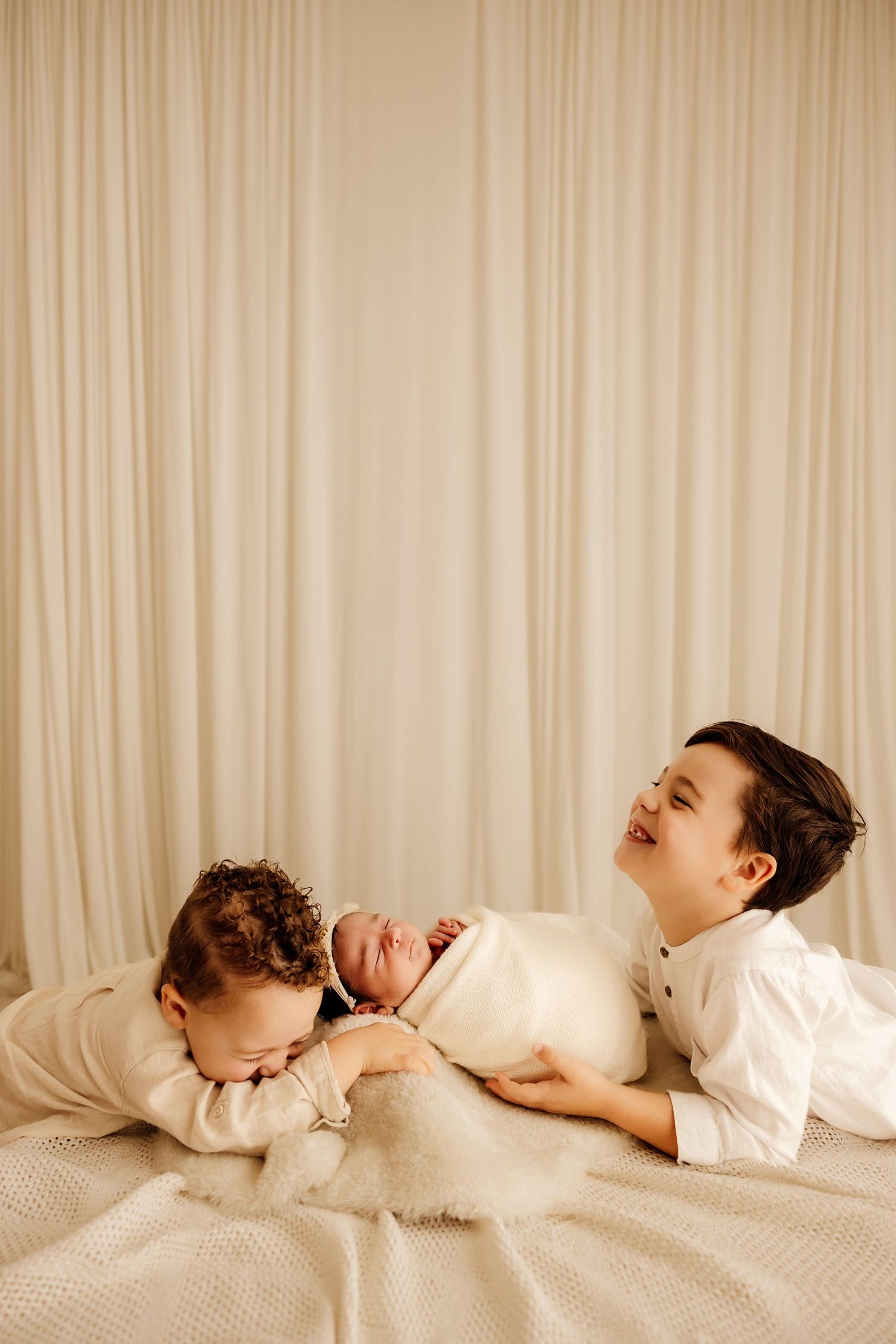 Two older siblings lying on a bed with a swaddled newborn baby, smiling and bonding in a soft, neutral-toned setting.