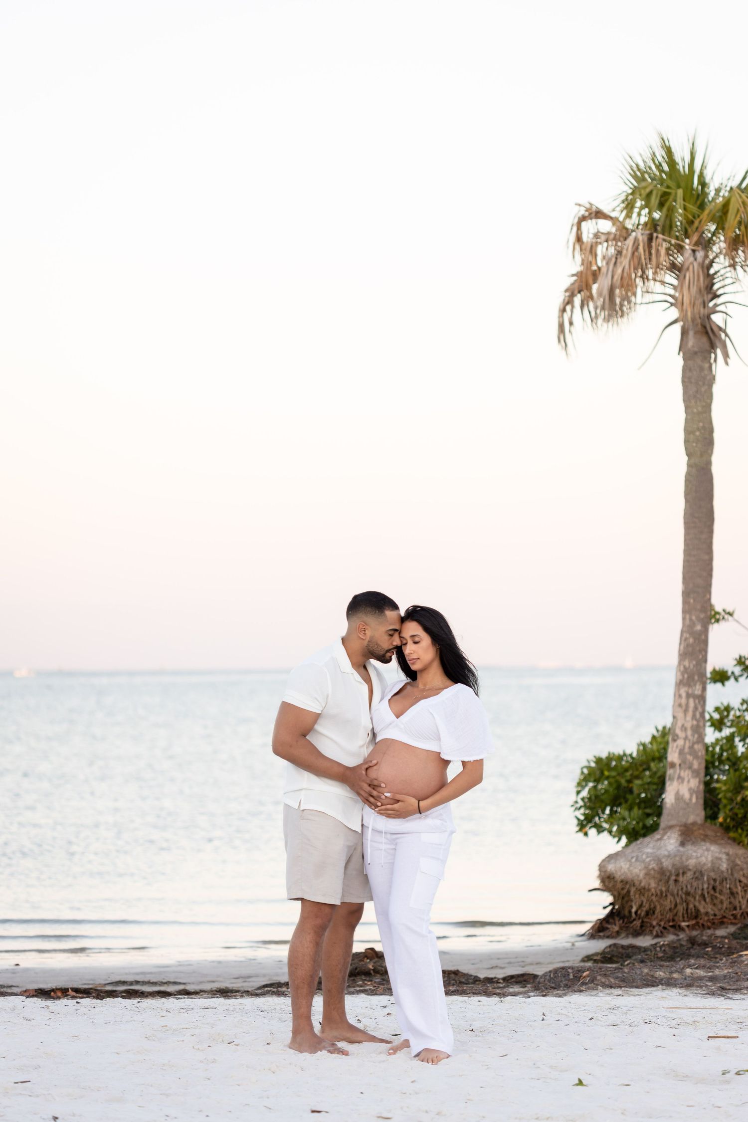 A romantic couple dressed in white embracing on a pristine beach with palm trees and ocean waves at sunset.