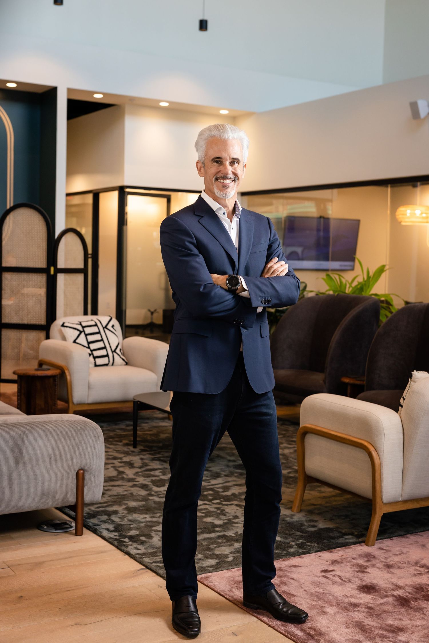 A confident middle-aged businessman in a navy suit stands with arms crossed, smiling in a modern lounge setting.