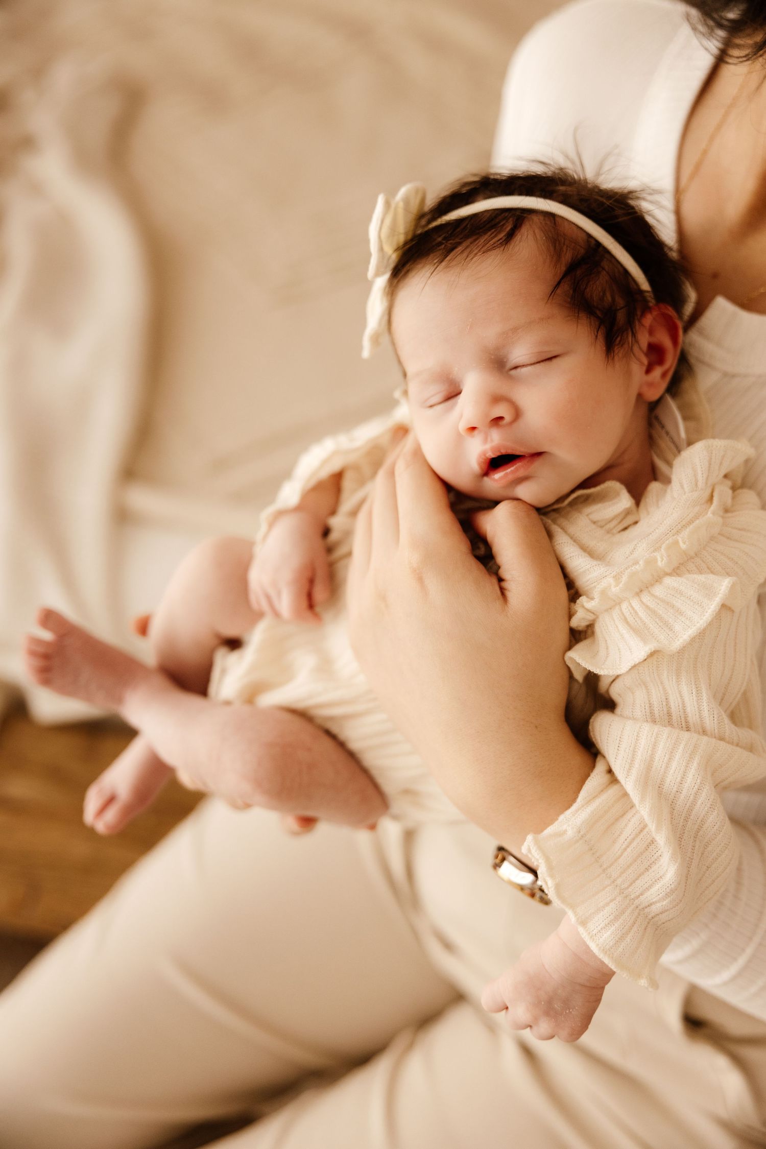 A newborn baby girl in a cream ruffled outfit and bow headband sleeps peacefully in her mother's arms.