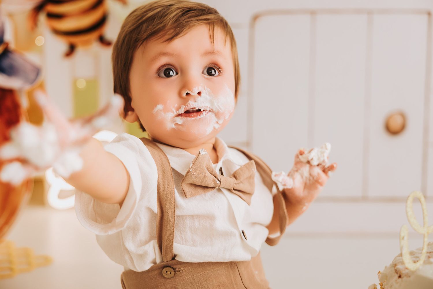 A toddler boy in a bow tie and suspenders smiles with cake smash frosting on his face and hands.