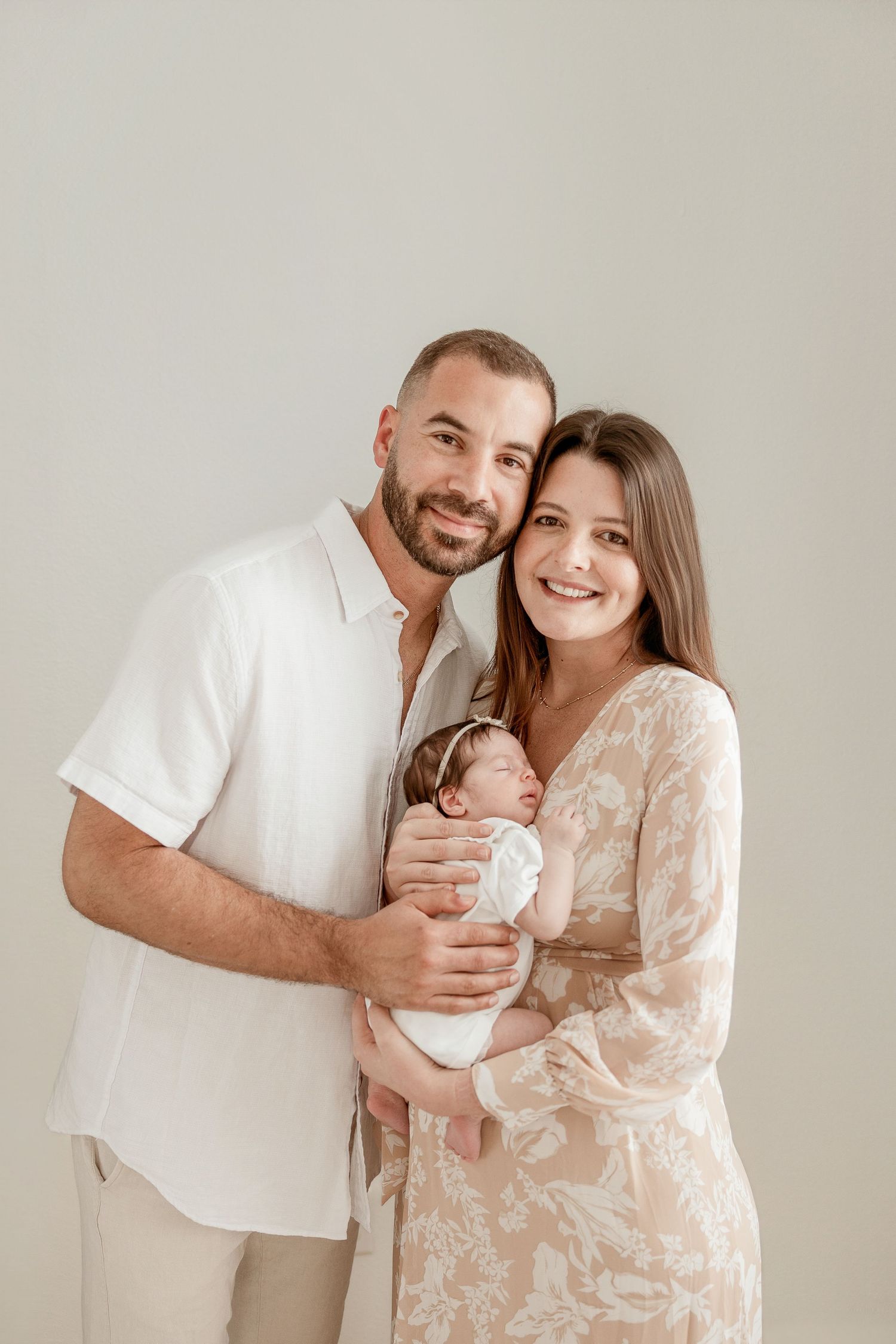 A smiling couple in neutral tones holds their swaddled newborn baby girl in a bright studio portrait.