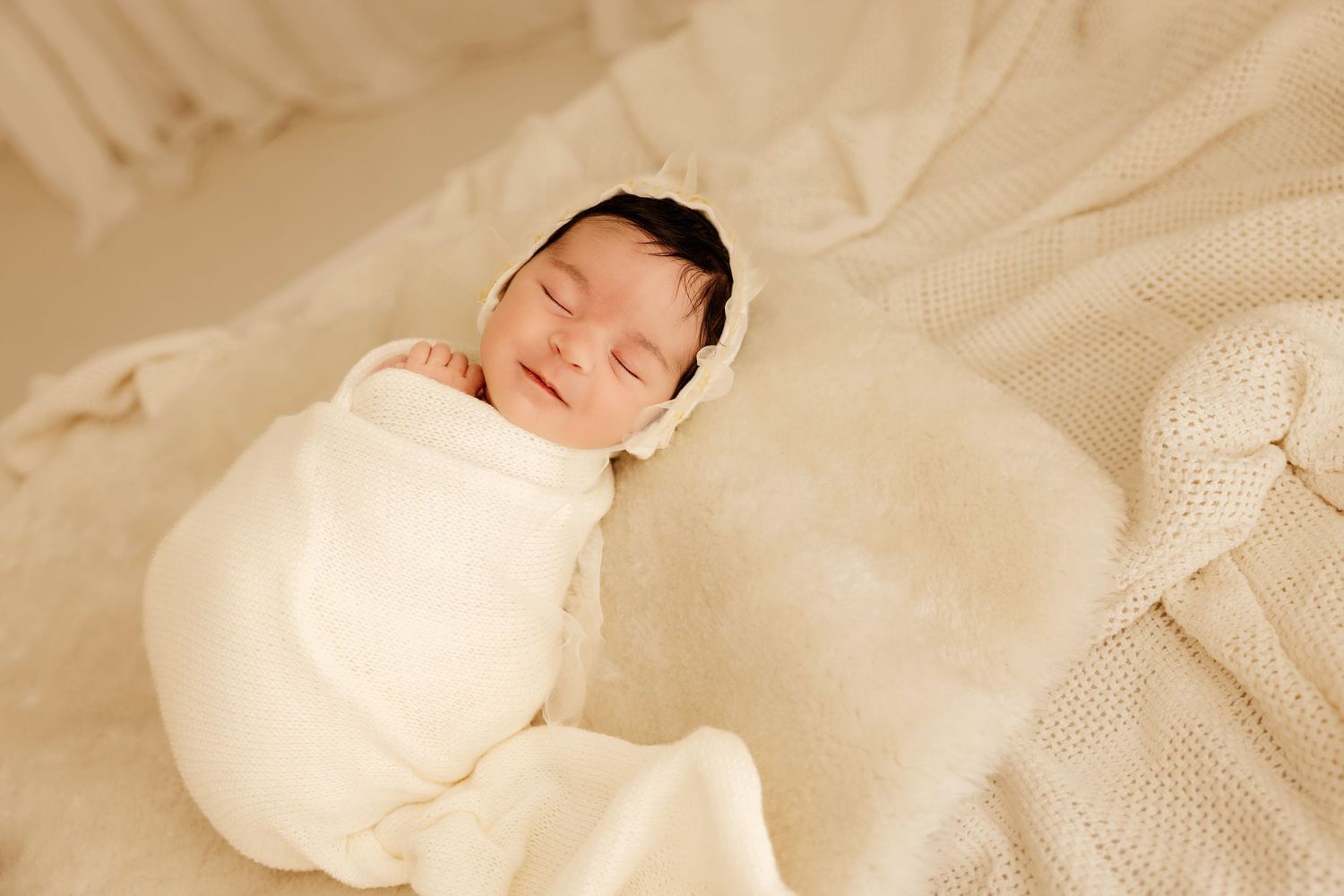 Newborn baby swaddled in white wrap and bonnet sleeping peacefully on a soft cream knit blanket.