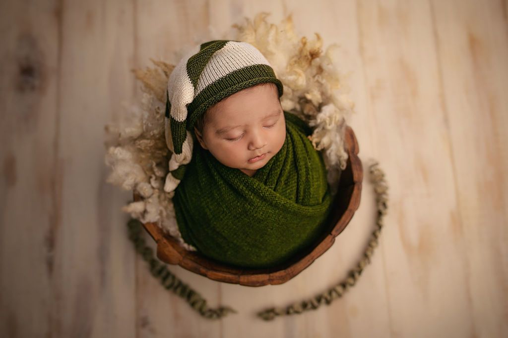 Sleeping newborn wrapped in green swaddle with matching knit hat nestled in wooden bowl with fuzzy decor.