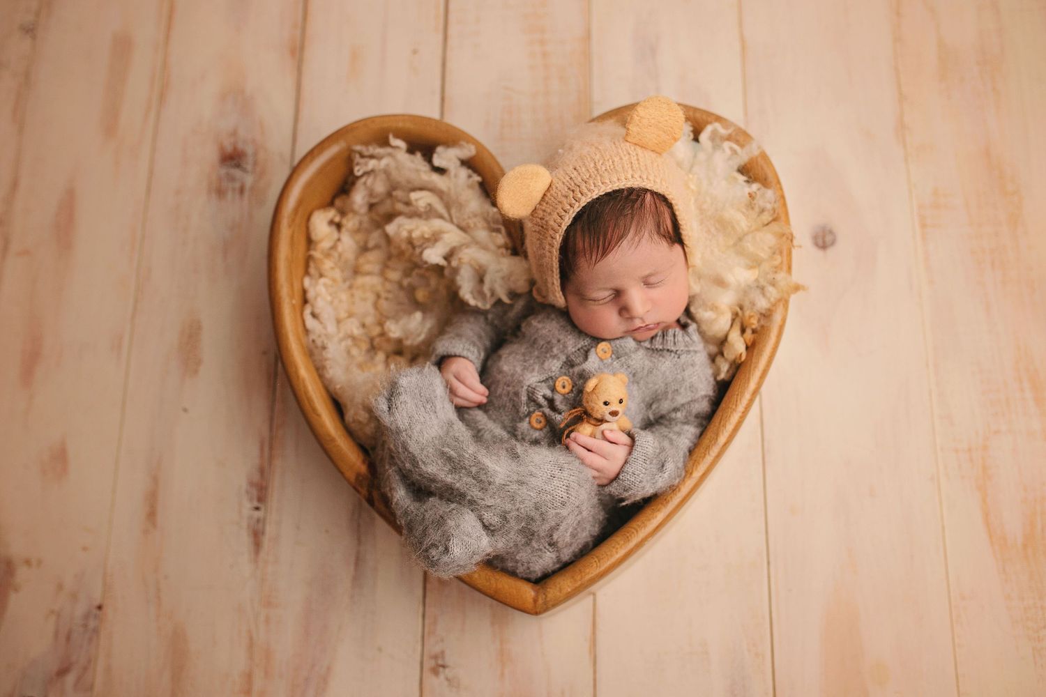 Newborn baby sleeping in heart-shaped wooden bowl wearing bear bonnet, holding tiny teddy bear on fluffy gray blanket.