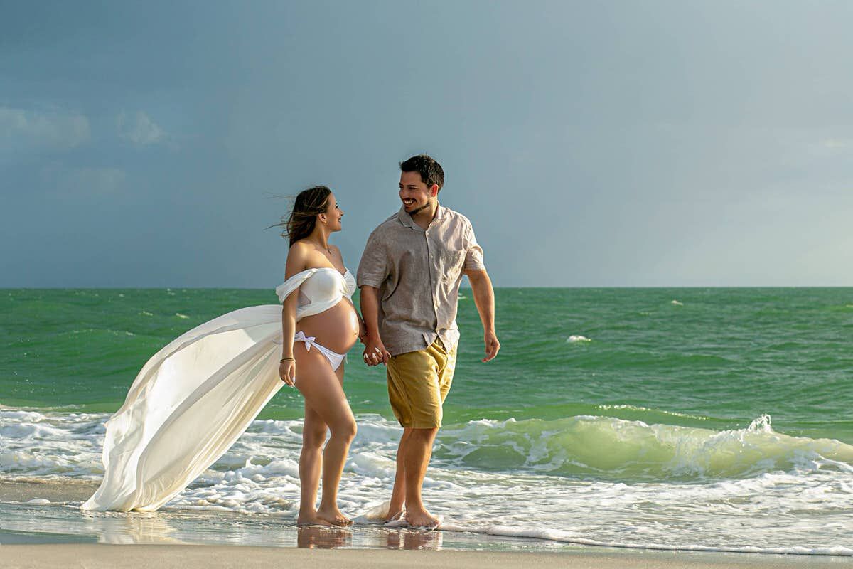 A couple stands together on a beach with turquoise waves and white sand as the woman holds a surfboard.