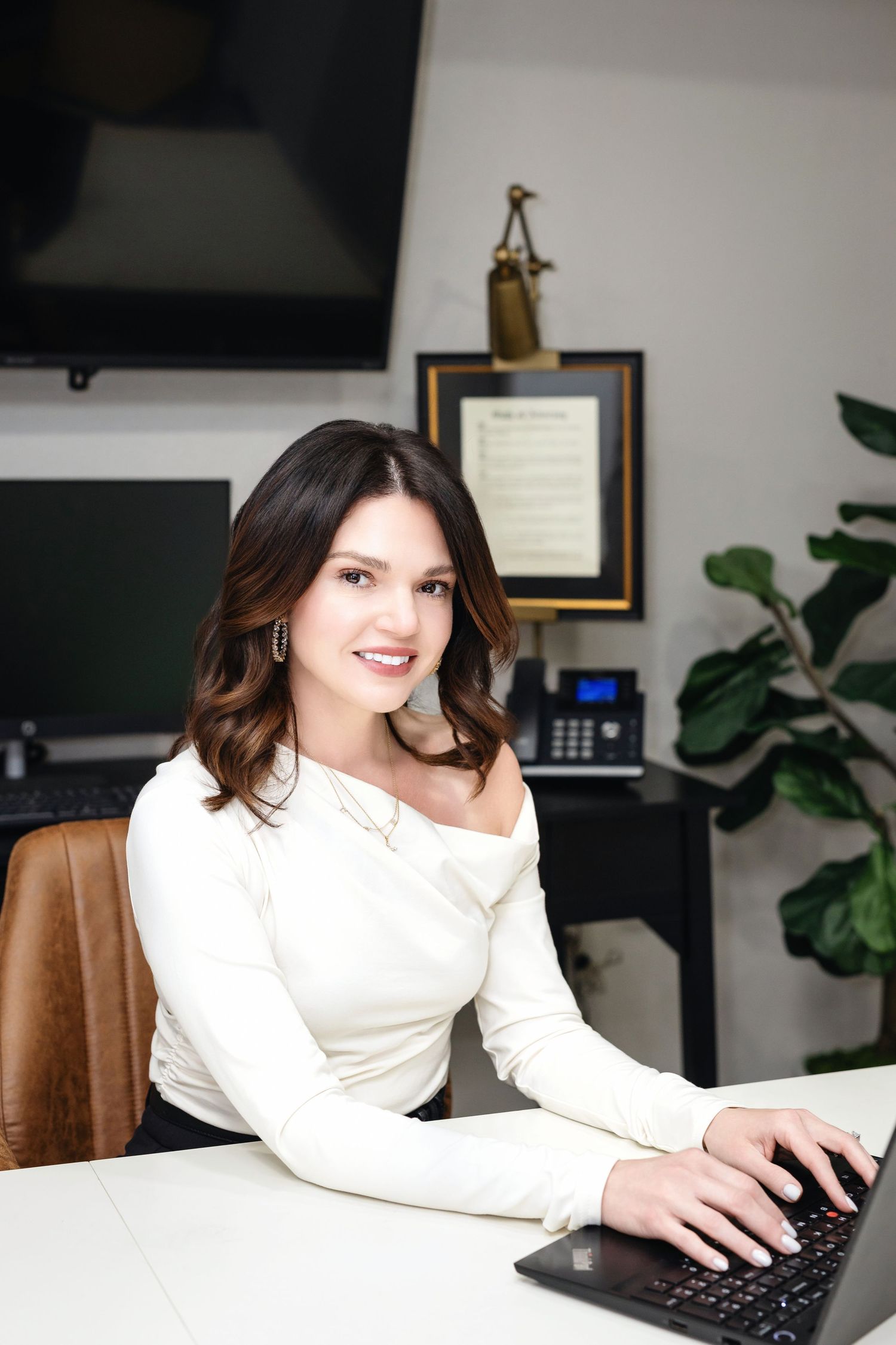 Smiling brunette woman in white off-shoulder top working on laptop at office desk with plant and framed art behind her.