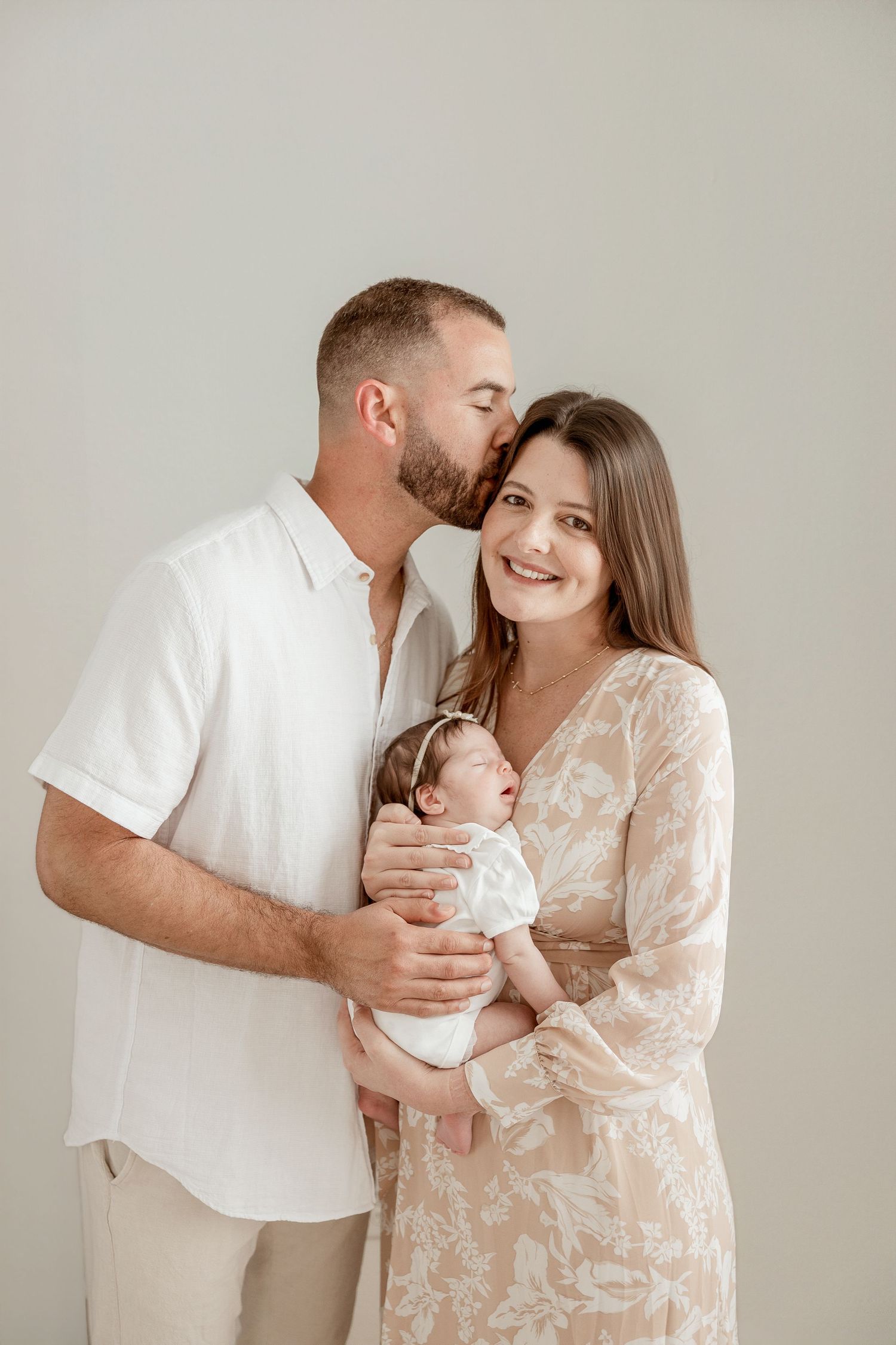 A couple in neutral tones holds their newborn baby, the man kissing the woman's cheek as she smiles warmly.