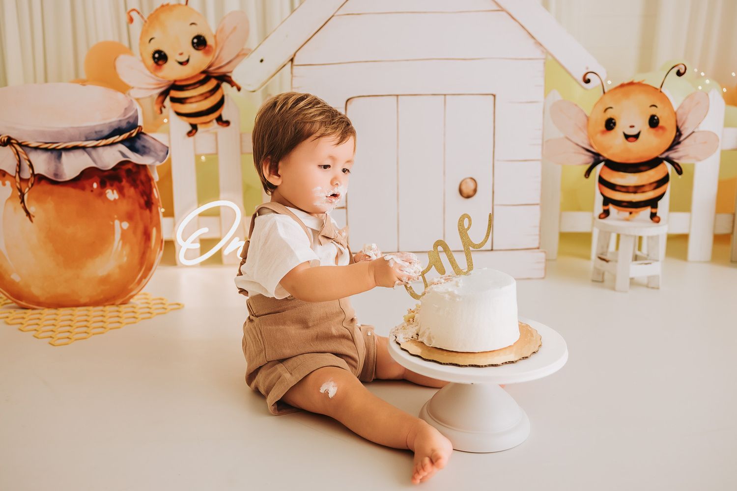 Baby boy in suspenders smashes first birthday cake surrounded by bee-themed decorations and honey jar props.