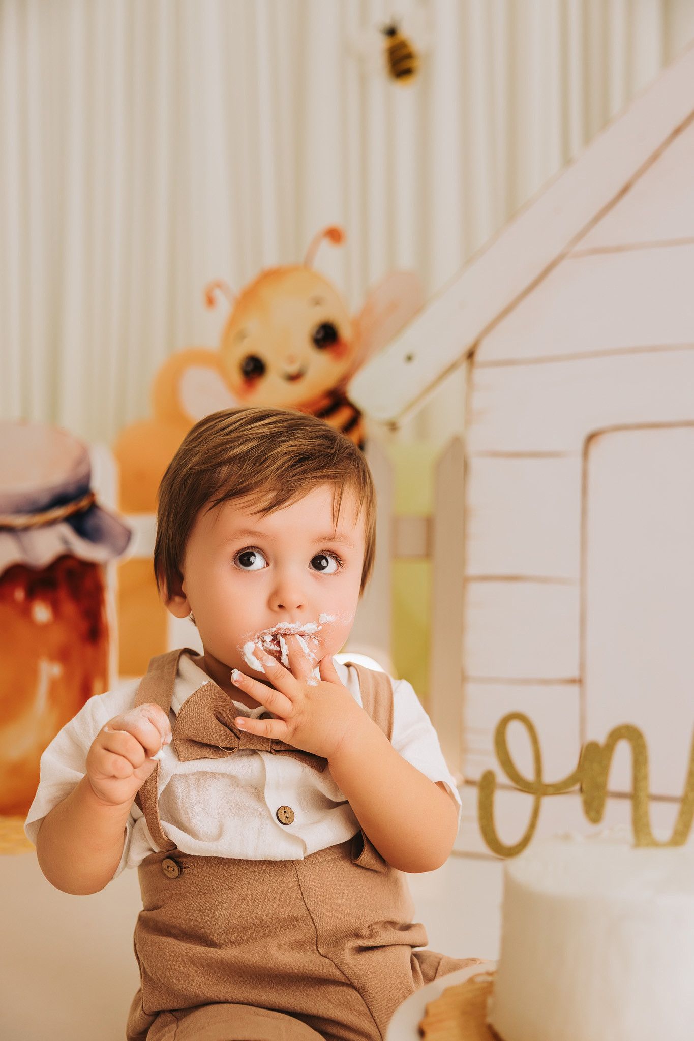 Toddler boy in brown overalls eating birthday cake at a bee-themed first birthday party.