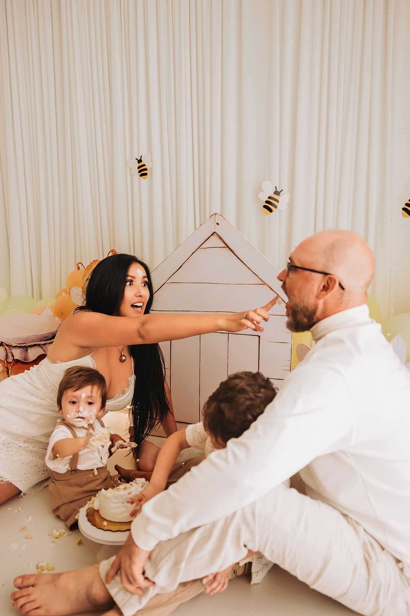 Family celebrating baby first birthday with bee-themed decor, cake smash, and cardboard house backdrop.