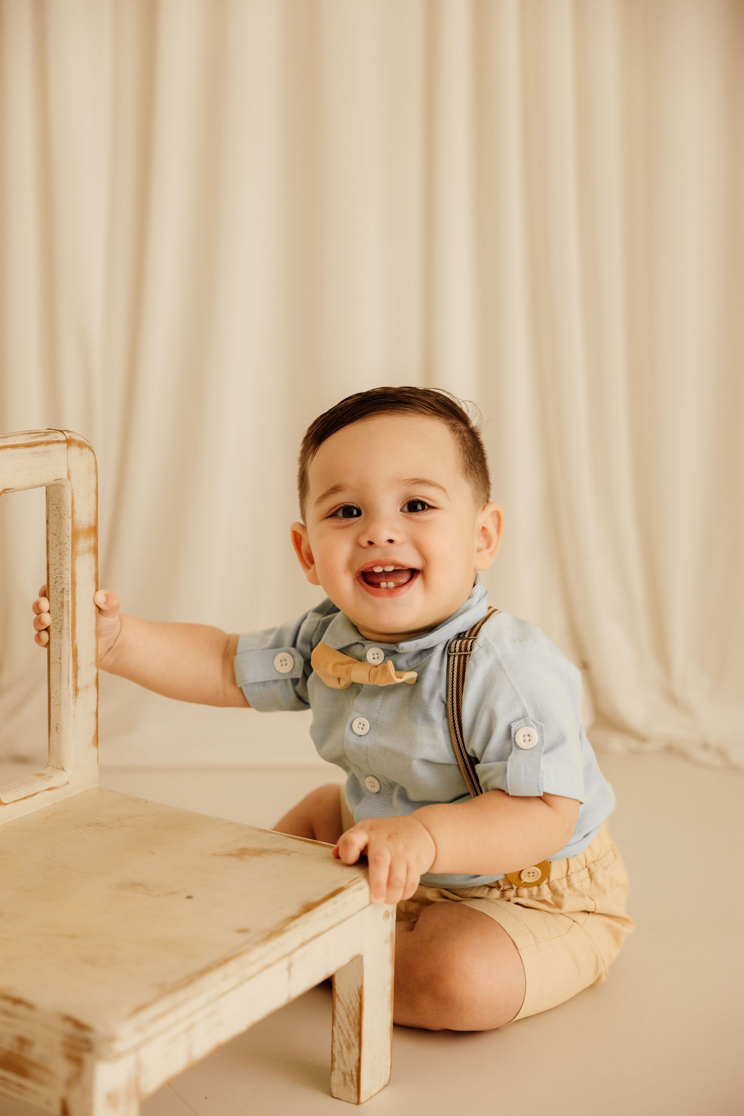 Smiling baby boy in blue shirt with suspenders and bow tie sits beside a rustic wooden table on a cream backdrop.