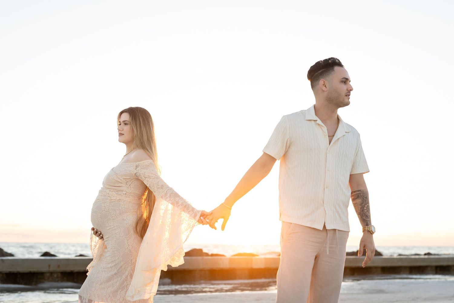 A couple in white outfits holds hands on a beach at sunset with warm golden light glowing behind them.