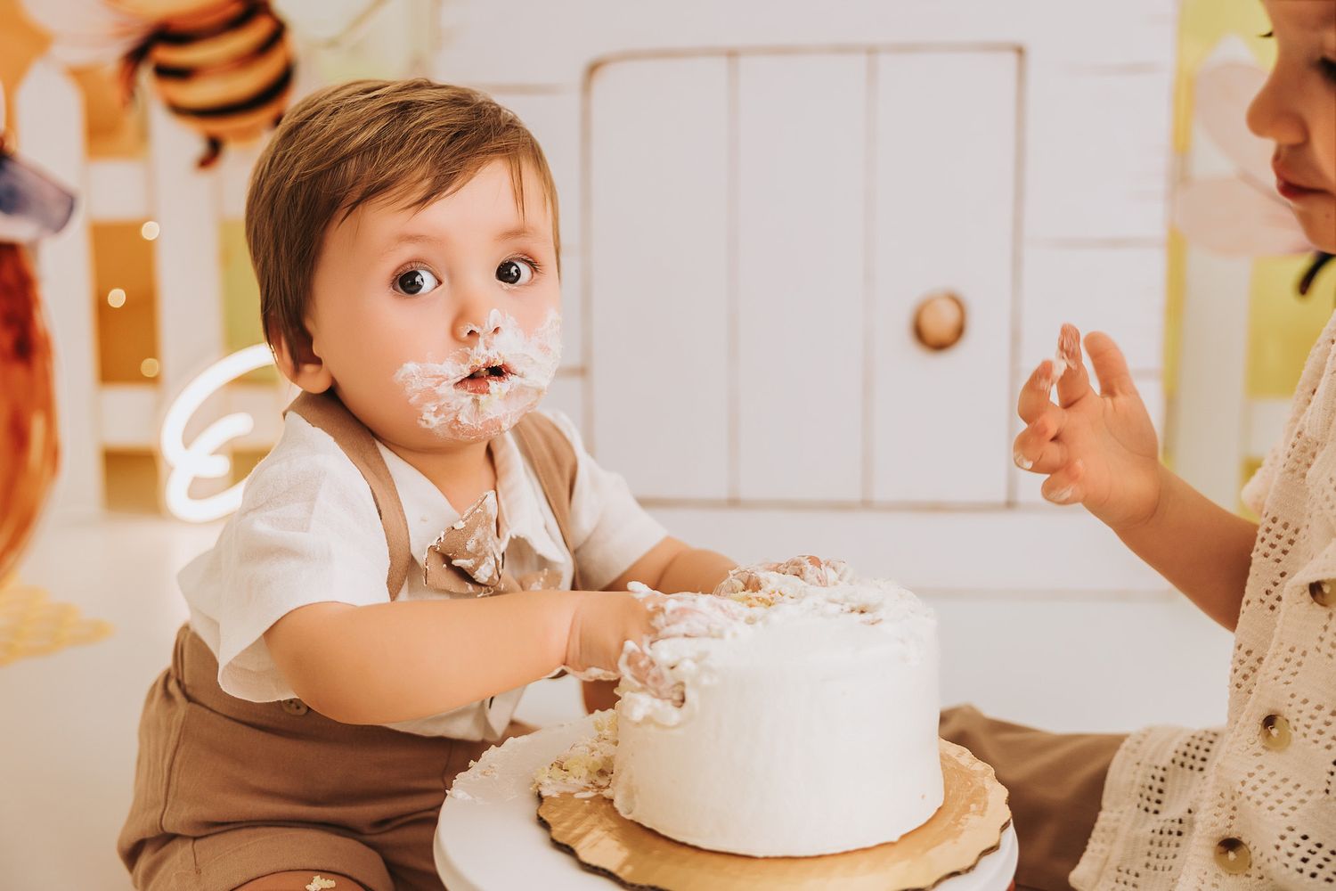 Baby boy smashing white cake at first birthday party, face covered in frosting, sitting at table.